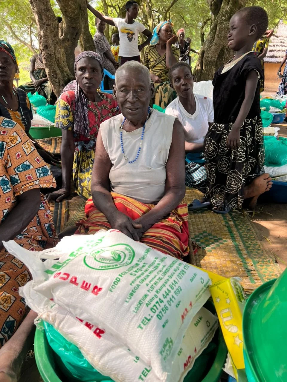 A group of people gathered outdoors under trees, with some sitting on mats and others standing. An elderly woman in the center is sitting with a bag of food in front of her, surrounded by women and children.