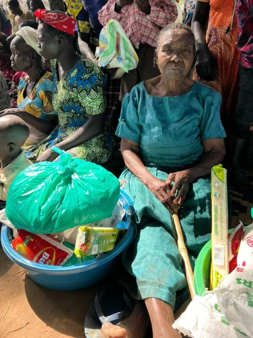 An elderly woman sitting on the ground with a walking stick, surrounded by people, with a large green bag and supplies in a blue basin nearby.
