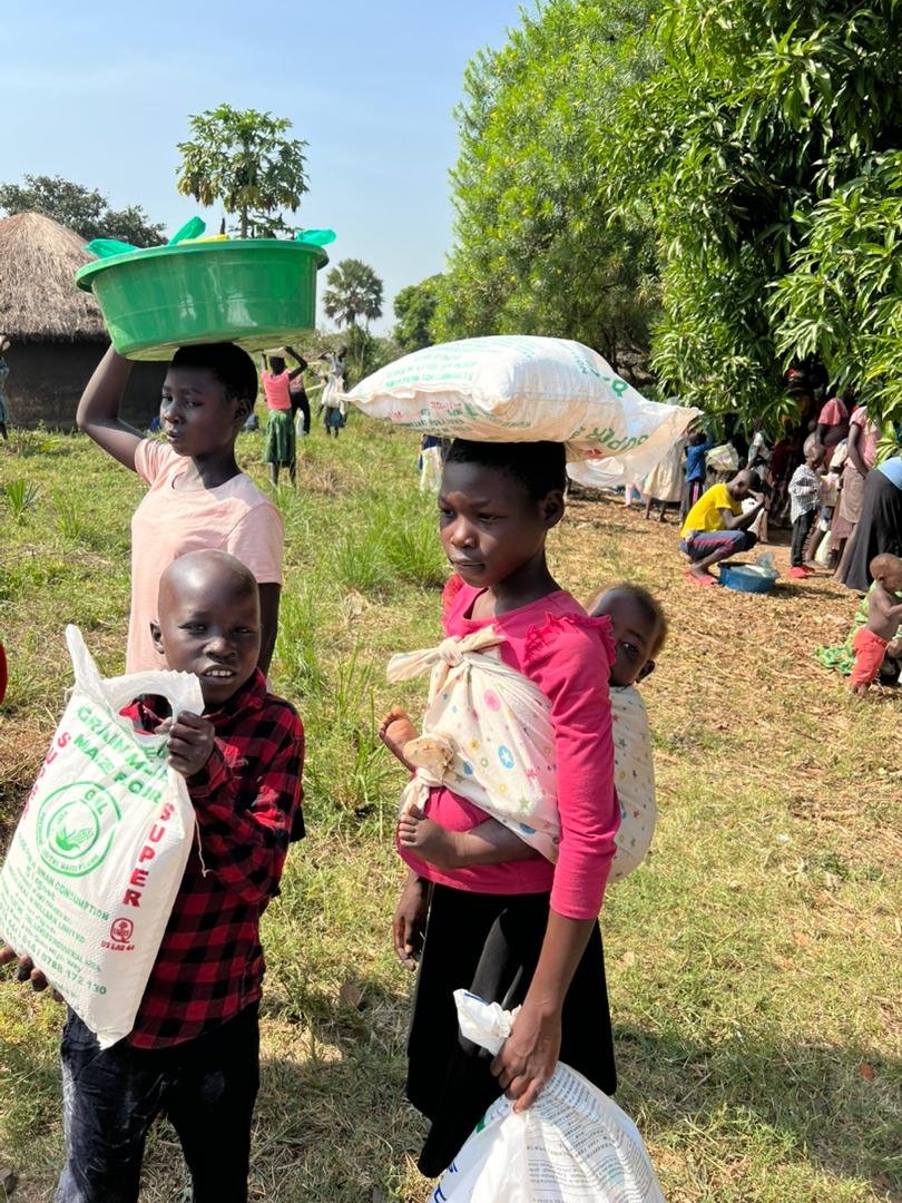 Group of children standing outdoors in a rural area, carrying bags and baskets, with trees and more children in the background.