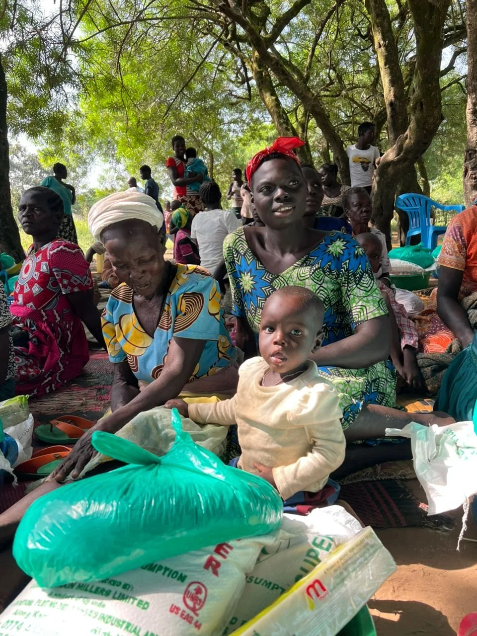 Group of African women and children gathered outdoors under big trees, sitting on mats with bags and supplies.