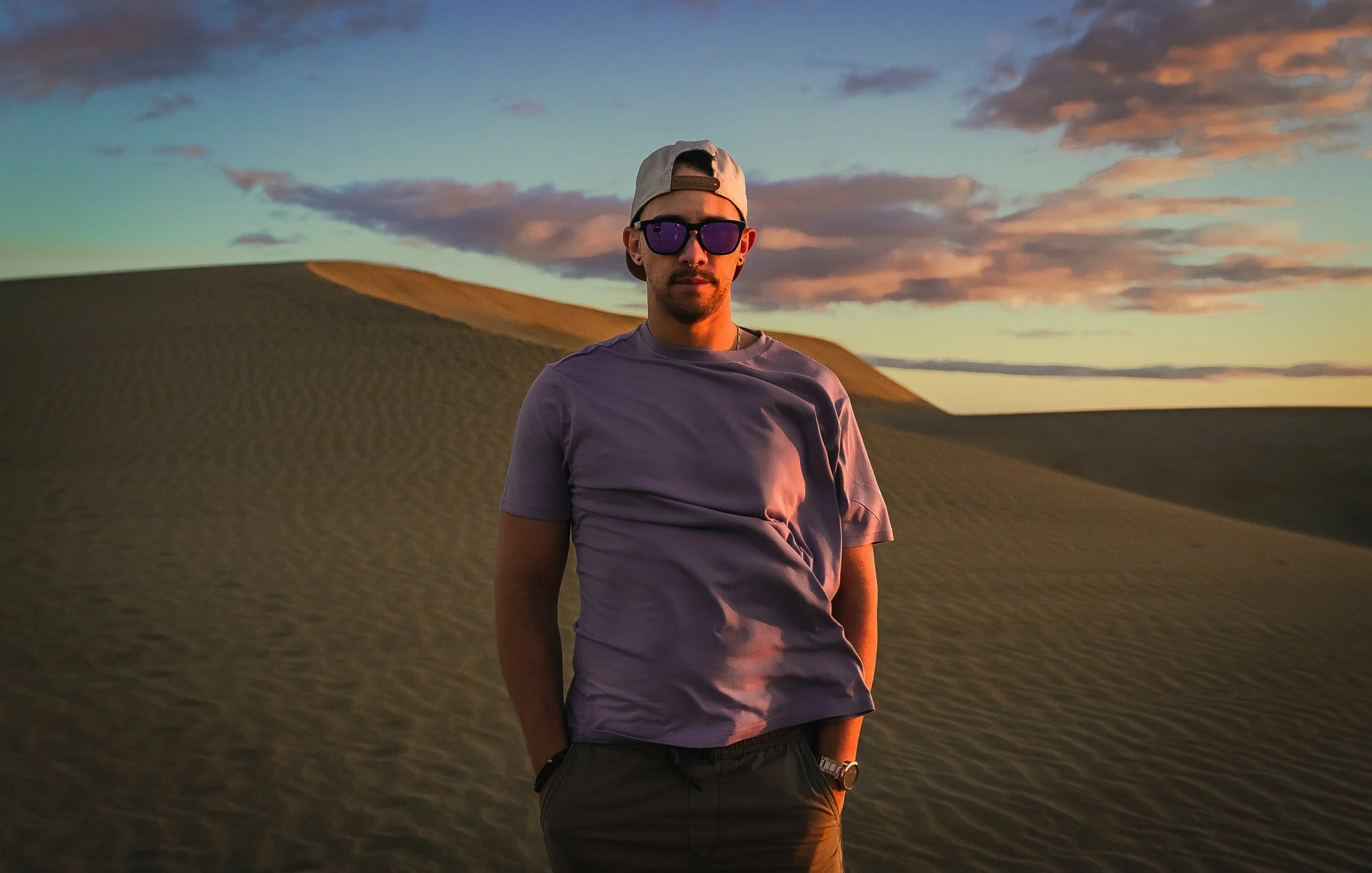 A man wearing sunglasses, a t-shirt, a white cap backward, and a watch stands in a desert with sand dunes under a colorful sunset sky.