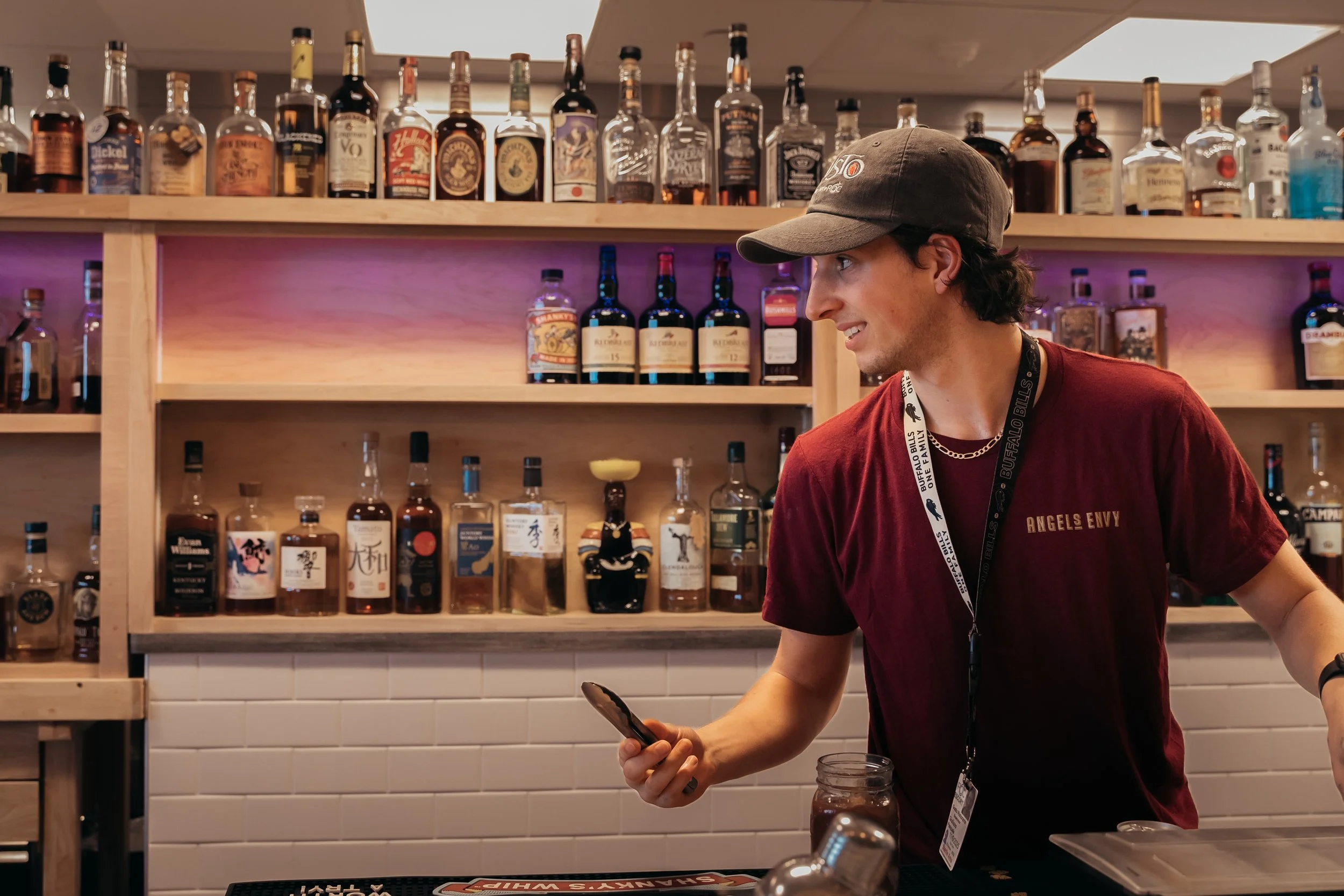 Young man wearing a baseball cap and a red 'Angels Envy' shirt behind a bar, holding a smartphone in his right hand, with a collection of liquor bottles on shelves in the background.