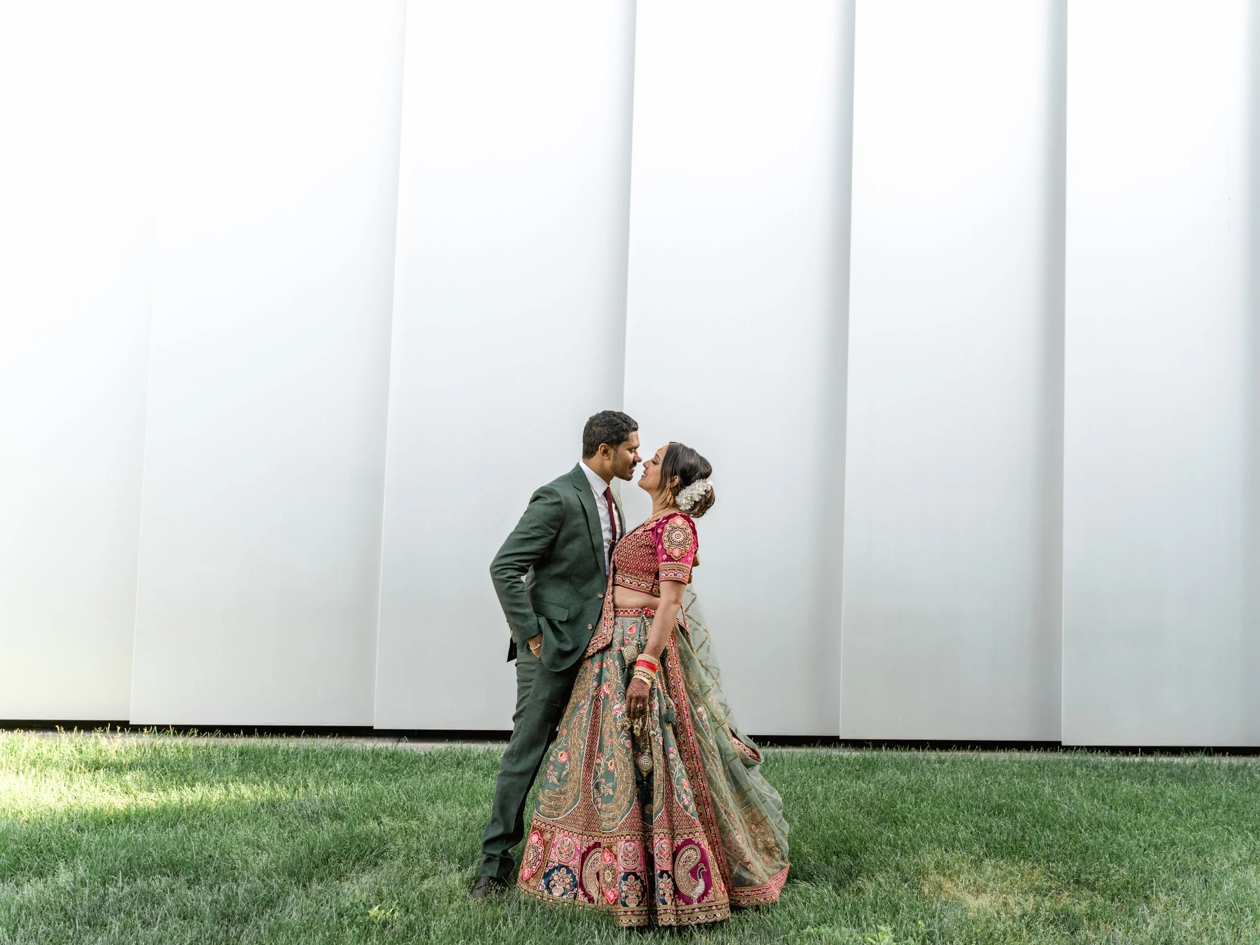 A couple dressed in traditional Indian wedding attire standing close on grass, with their faces almost touching, in front of a modern, white architectural backdrop.