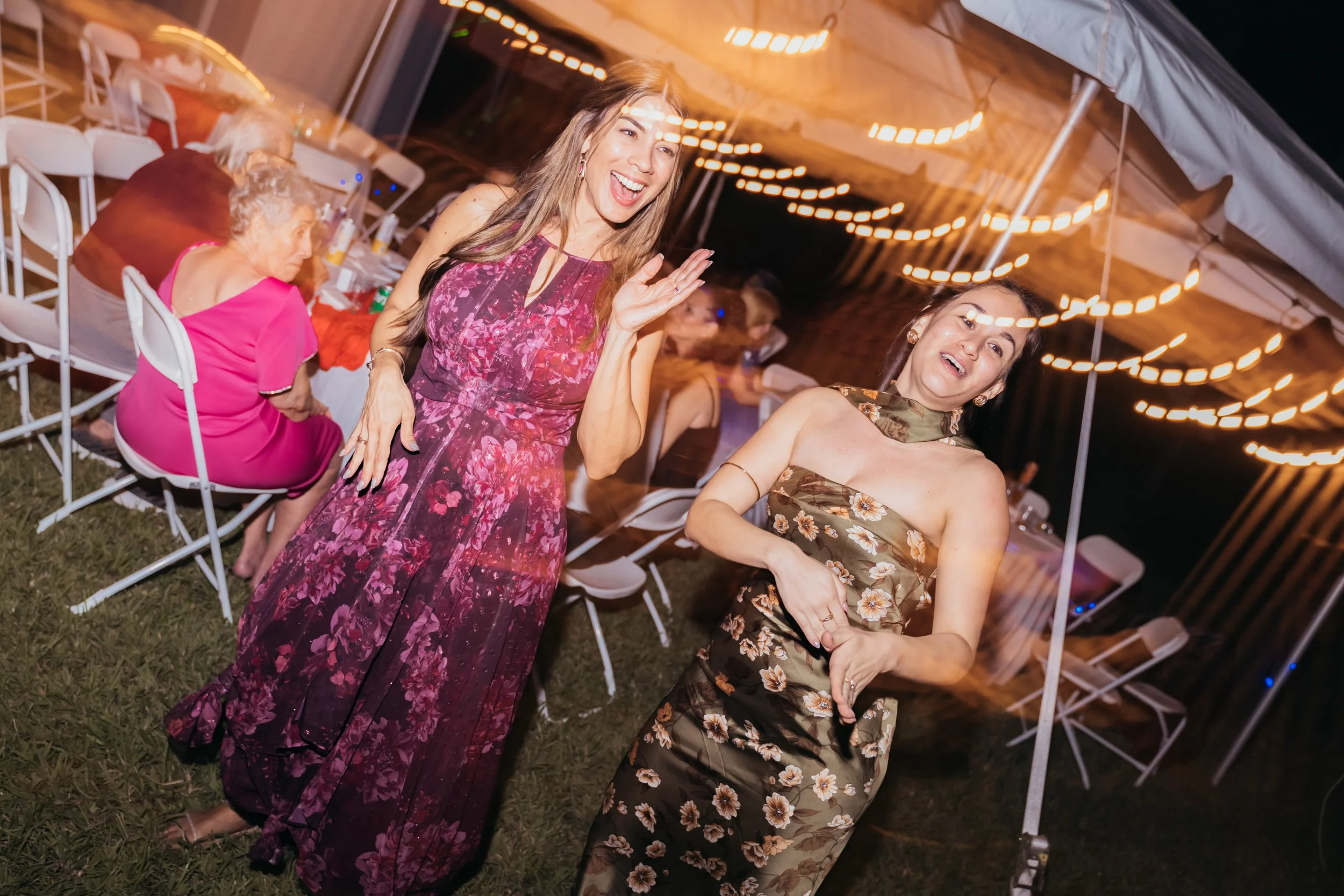 Two women dancing and smiling at an outdoor party, with tables and other guests in the background under string lights at night.