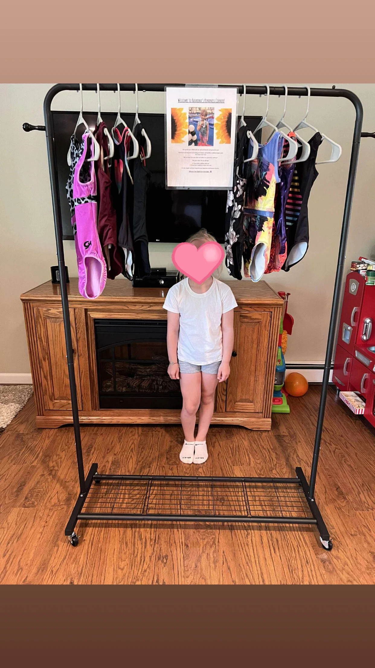 A young girl standing in front of a clothing rack with swimwear, in a room with wooden floors and furniture, with a television and toys in the background.