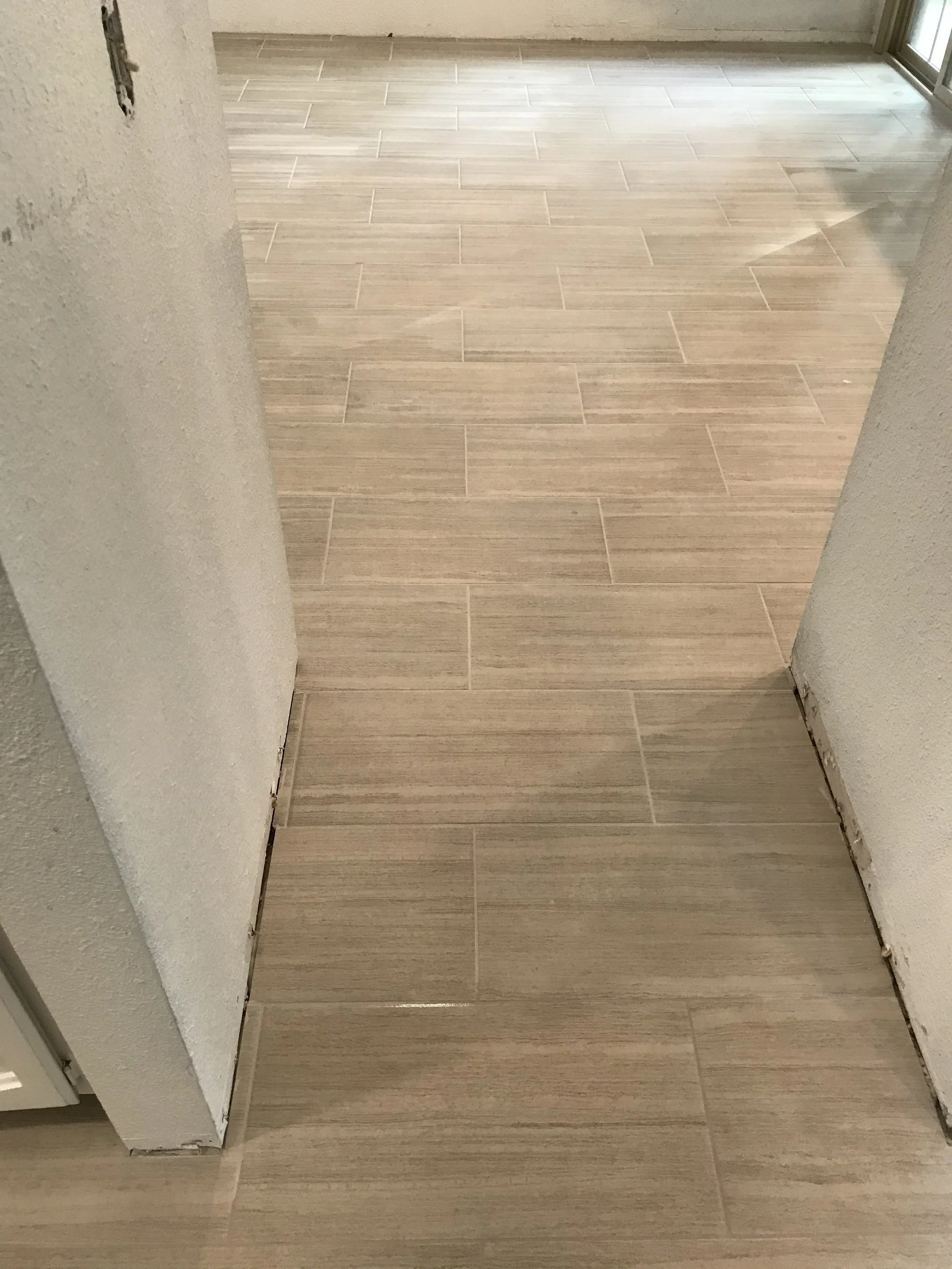 Interior of a room under renovation with light wooden floor tiles, an unfinished wall with visible damage, and a sliding glass door.