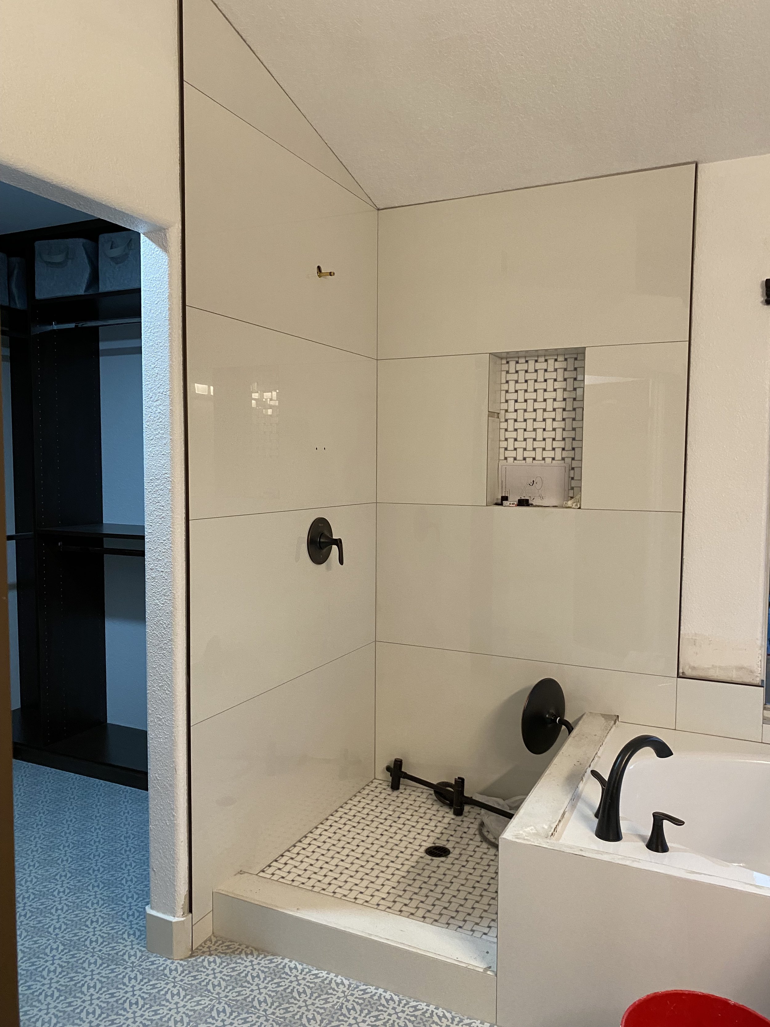 Bathroom shower area with beige tiles, a small mosaic tiled niche for toiletries, a black showerhead, and a white vessel sink with black fixtures on the right. To the left, a black shelf with storage bins is visible.
