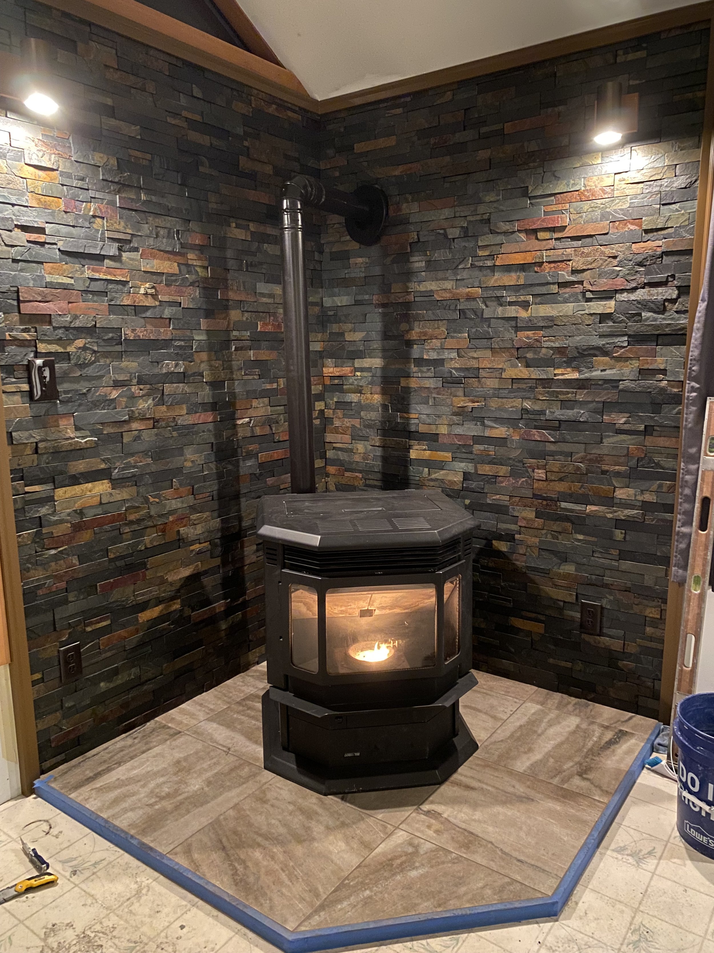 A corner of a room with a wood stove that has a fire inside, positioned on a tiled floor. The walls are covered with dark, multicolored stacked stone. There is a shadow on the wall cast by the stove and pipe, and a blue tape outline on the floor surr