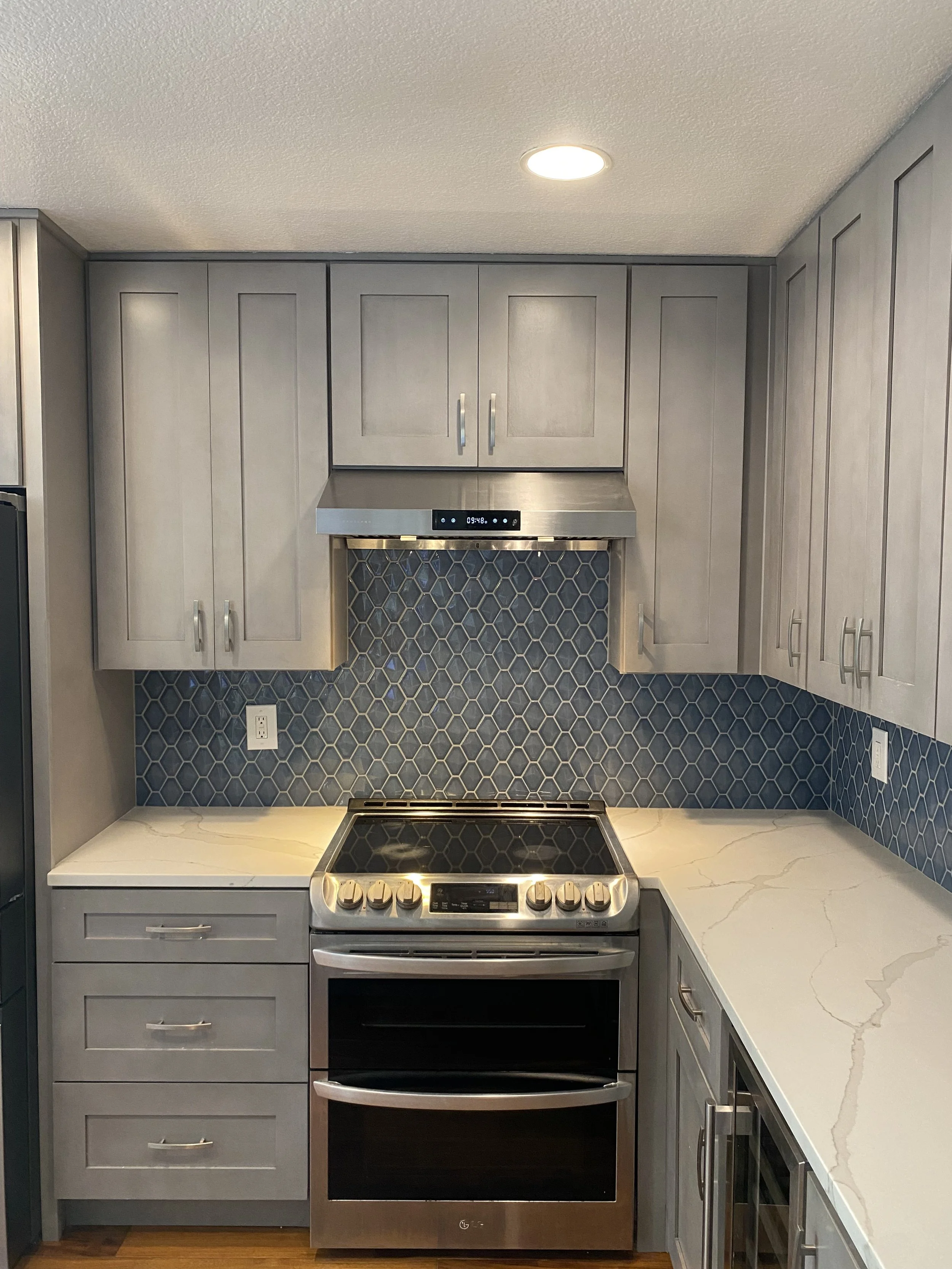 Kitchen with gray cabinets, stainless steel oven and range, white marble countertop, and a blue hexagonal tile backsplash.