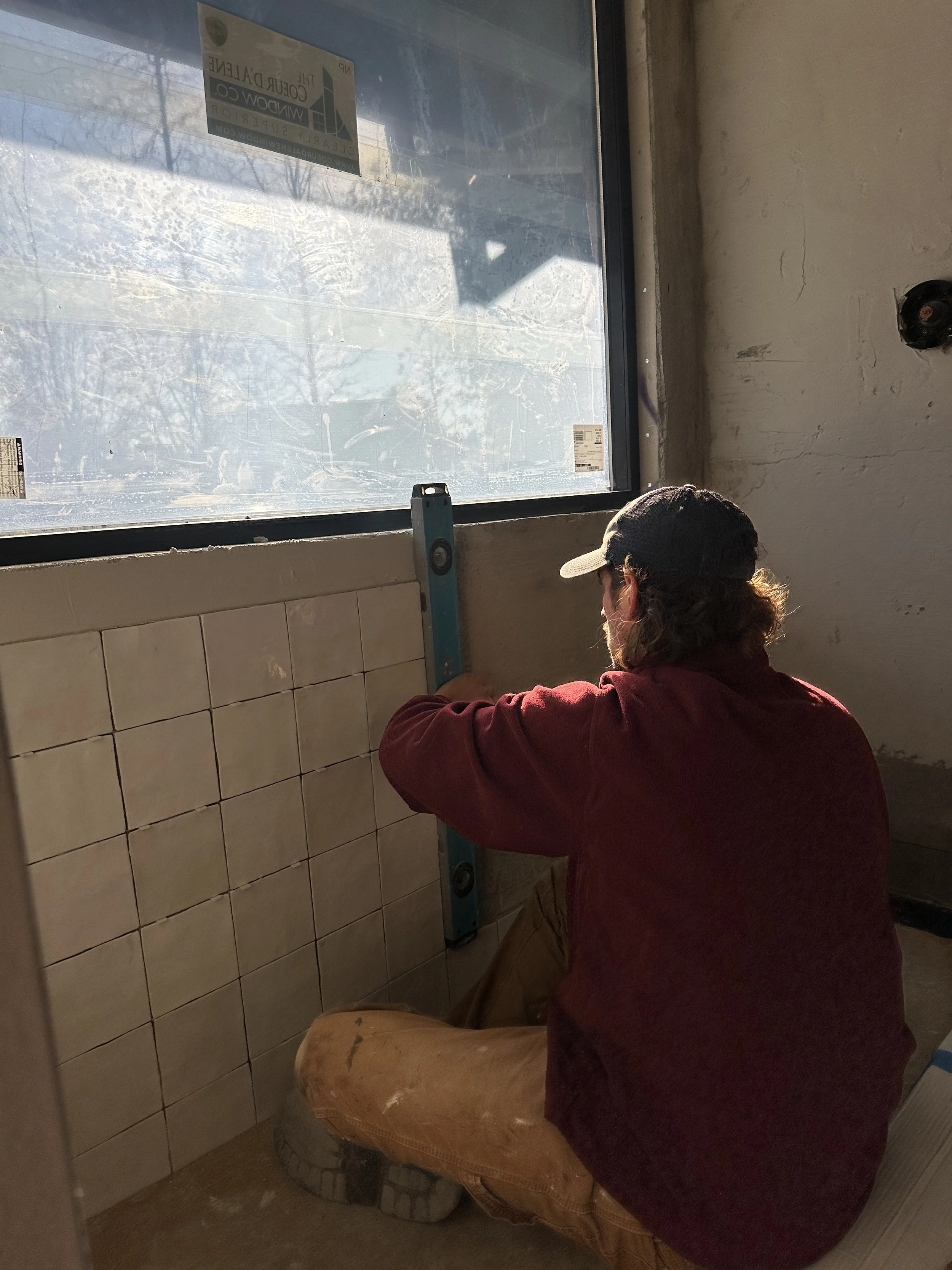 A man in a baseball cap and red shirt kneels on the floor, measuring the wall near a large window with a level tool. The room has beige tiles on the wall and a concrete floor.