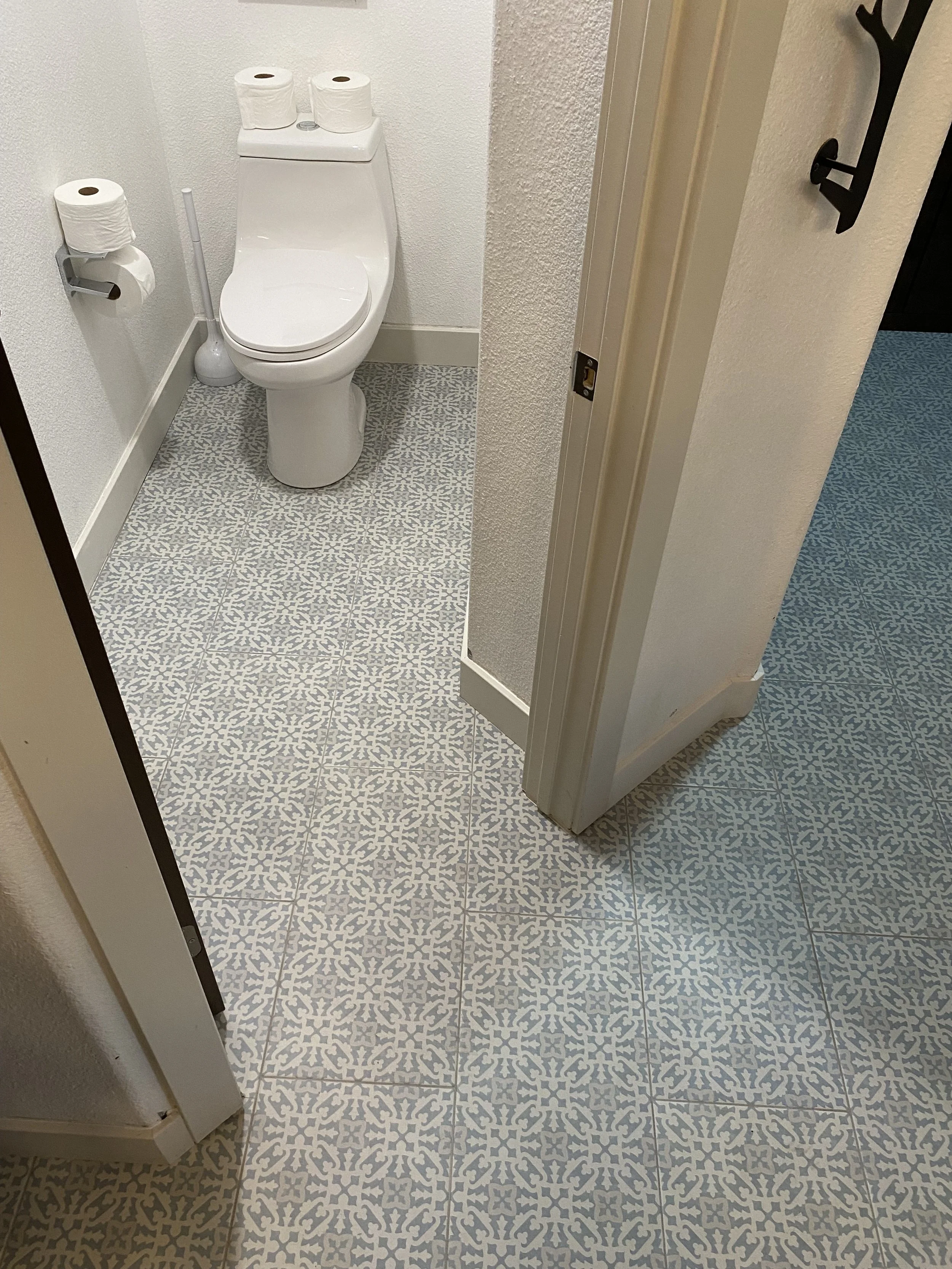 A small bathroom with a white toilet and patterned tile flooring. There are two rolls of toilet paper on top of the toilet tank, a toilet brush to the left, and a toilet paper holder with two rolls attached to the wall to the left. The bathroom door 