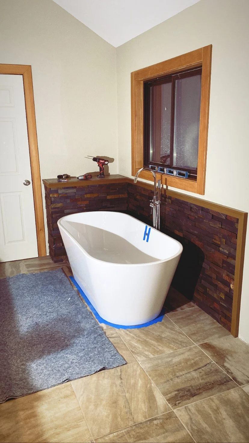 A bathroom with a freestanding white bathtub, a window with wooden trim, a dark brick half wall, and a shelf with tools including a drill and level.