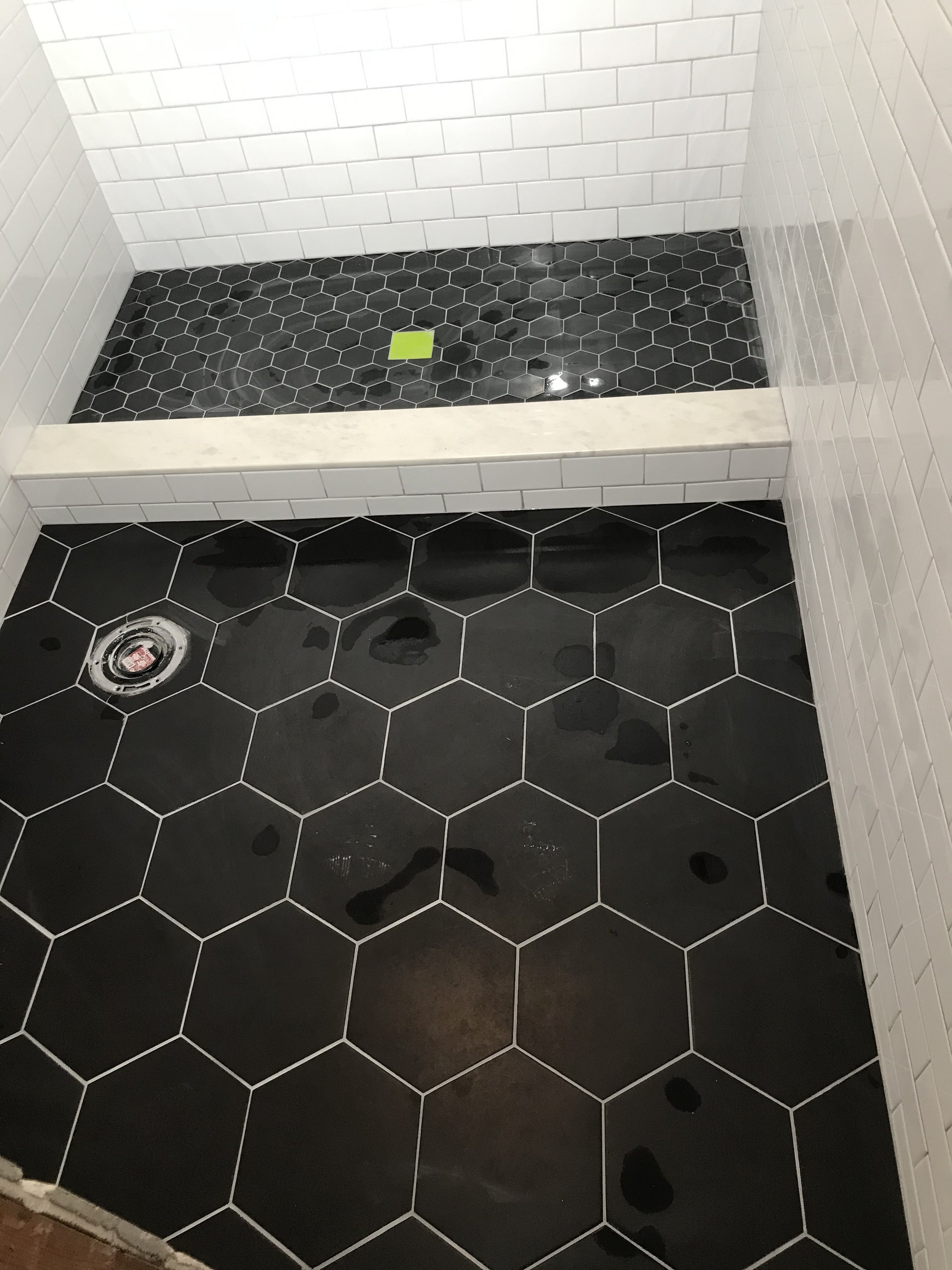 A corner of a bathroom shower with black hexagonal floor tiles, white rectangular wall tiles, and a white marble threshold. There are water spots on the floor and a drain in the lower left corner.
