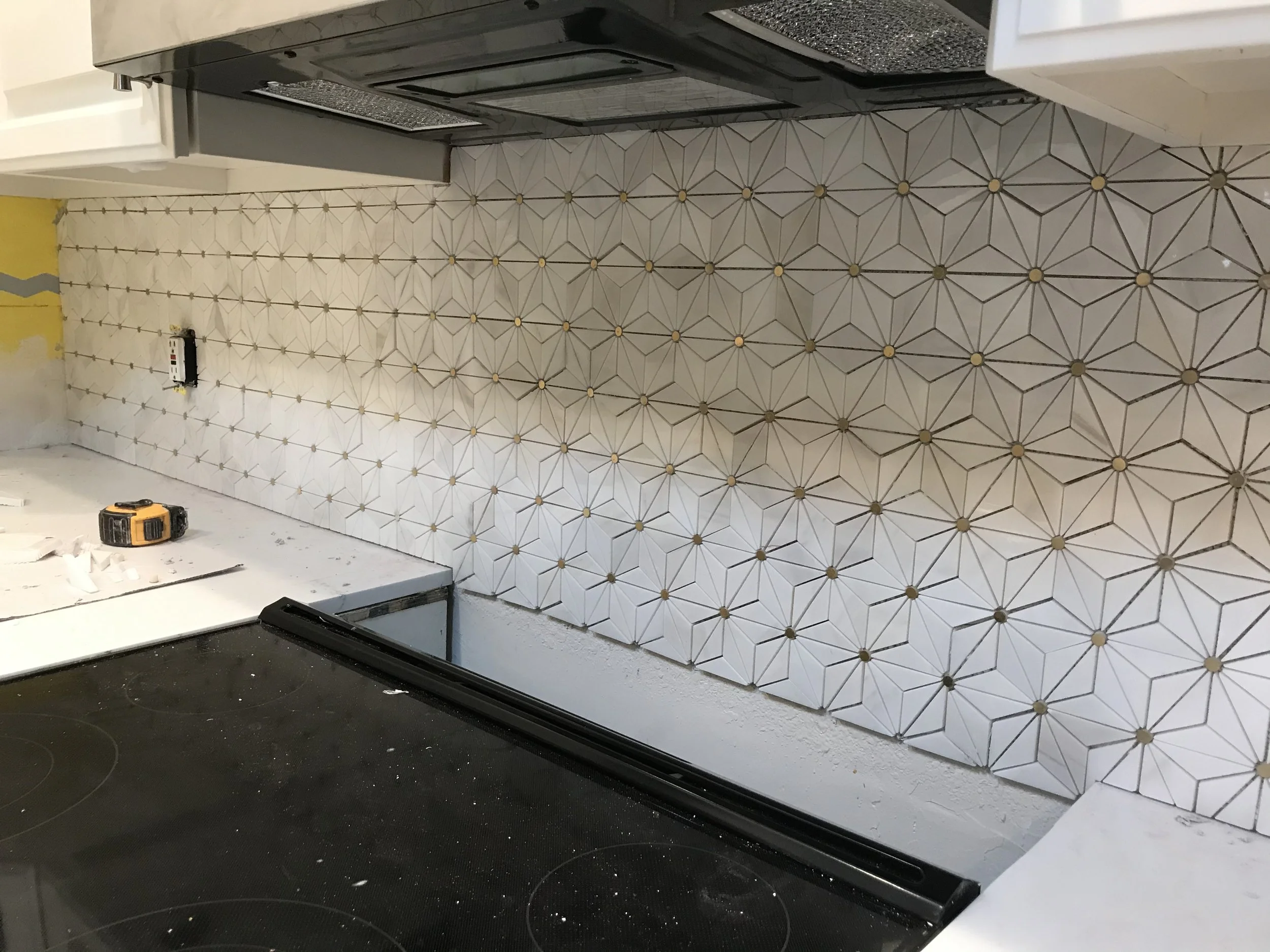 A kitchen wall with white tiles featuring geometric patterns and small round accents. Part of a black oven and a white cabinet are visible, with tools and a drill on the black countertop.