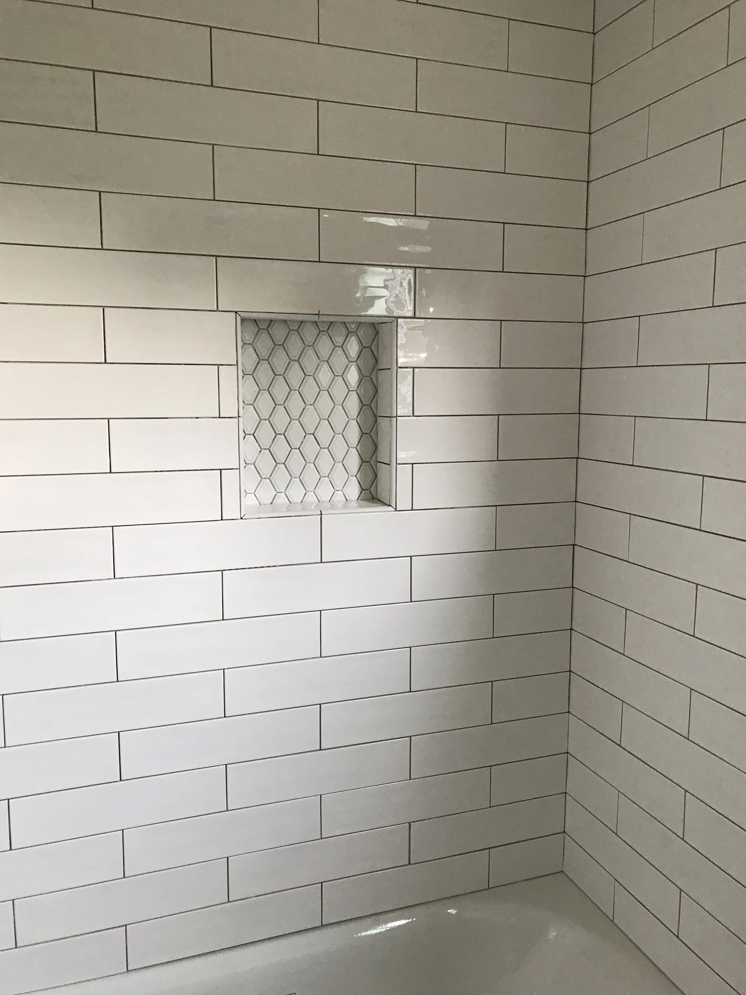 Bathroom wall with horizontal white subway tiles, a built-in niche with geometric white tile pattern, a small white ledge, and a white bathtub at the bottom."