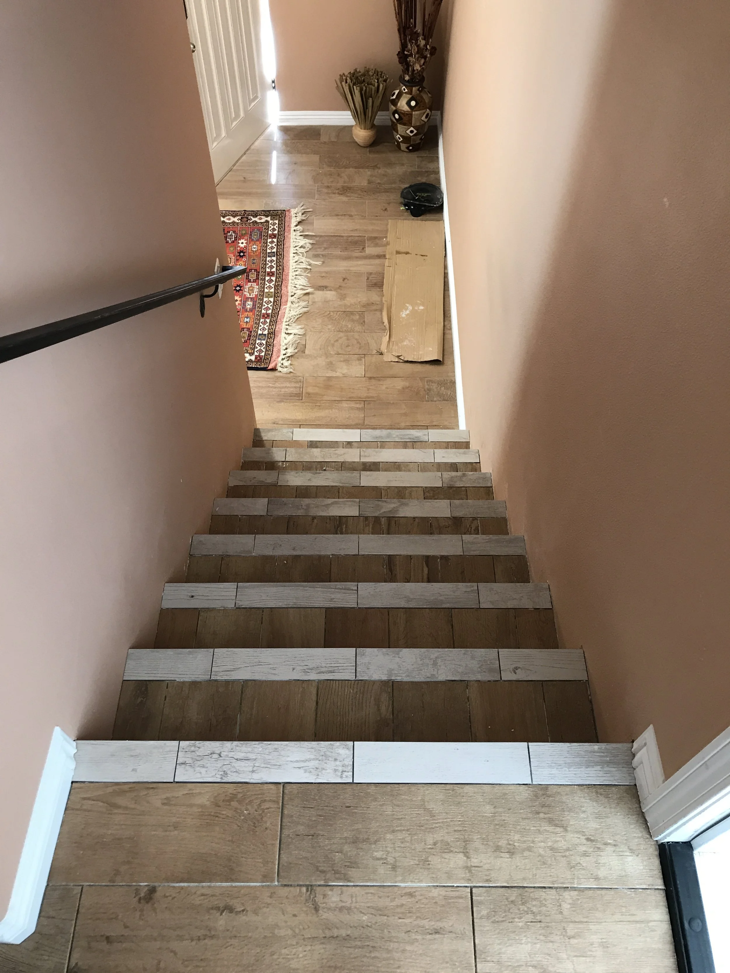 View from top of staircase showing hardwood steps, transitioning from darker to lighter wood, leading down to a hallway with a beige wall, a small decorative rug, a cardboard box, a black object, and vases with dried plants near a door.