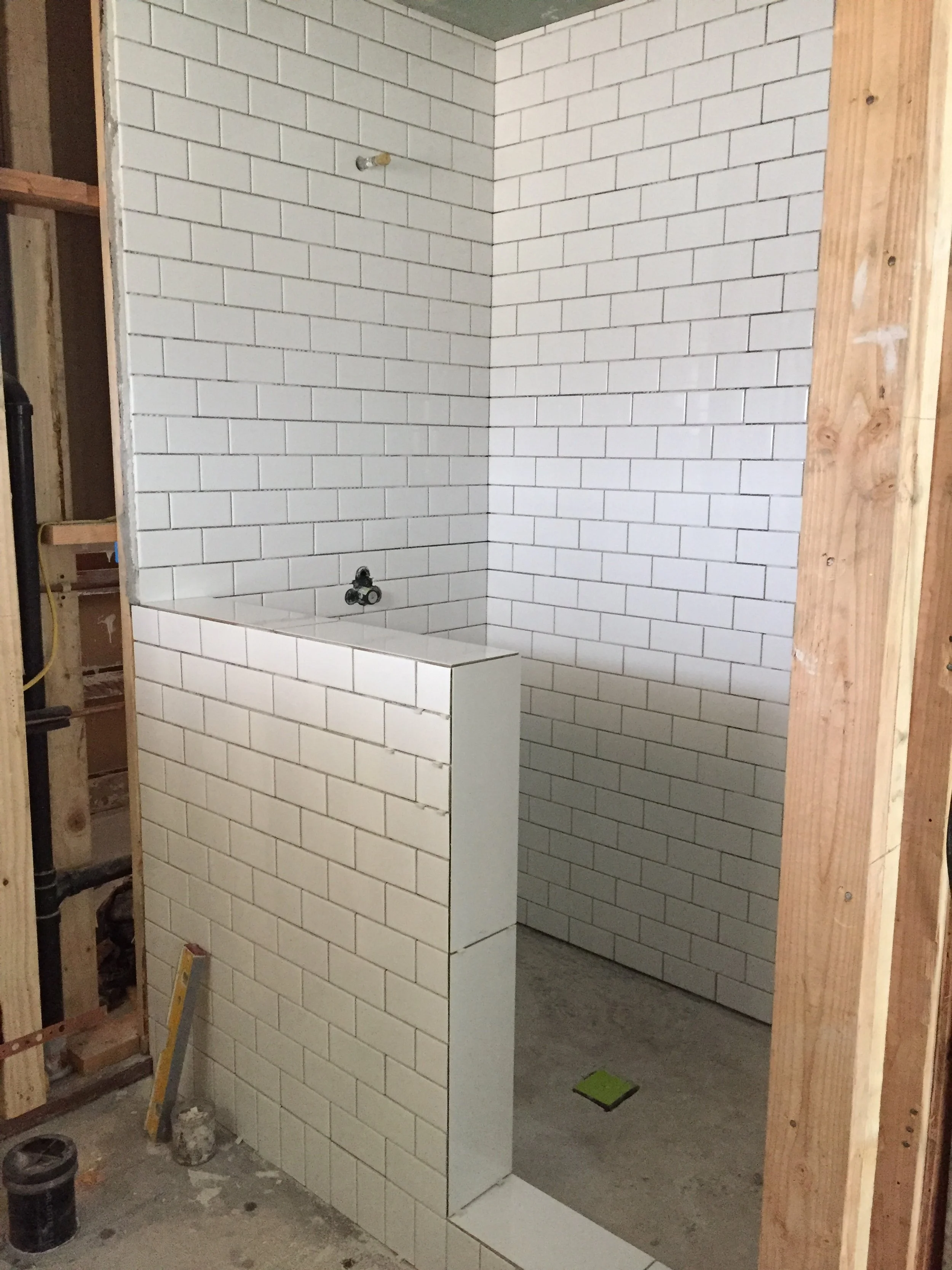 Construction site showing tiled wall with small white bricks, a corner structure with large black faucet fixtures, and wooden framing on the sides.