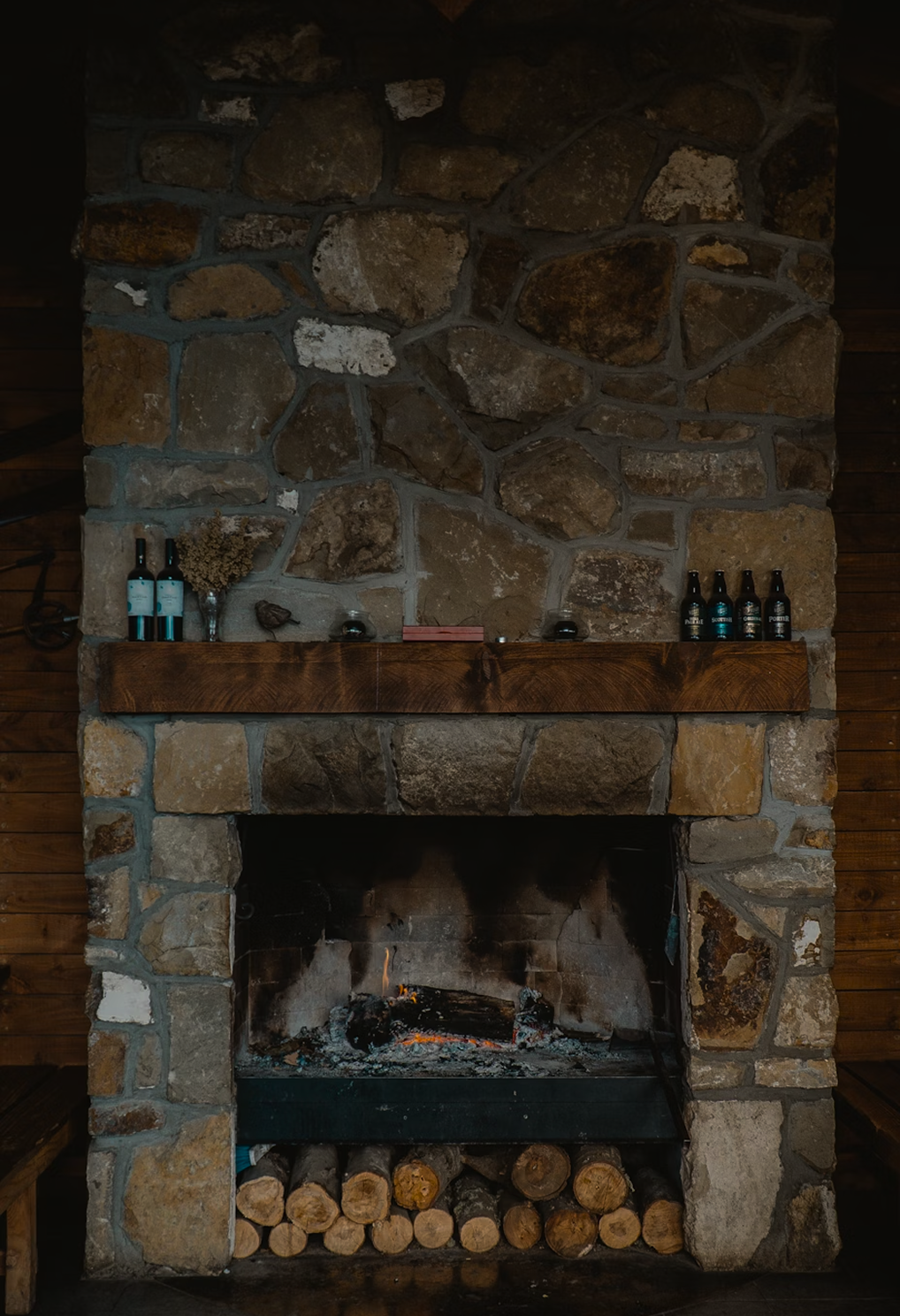 A rustic stone fireplace with a wood mantel, with three wine bottles and a glass with dried flowers on the mantel, and logs stored underneath.