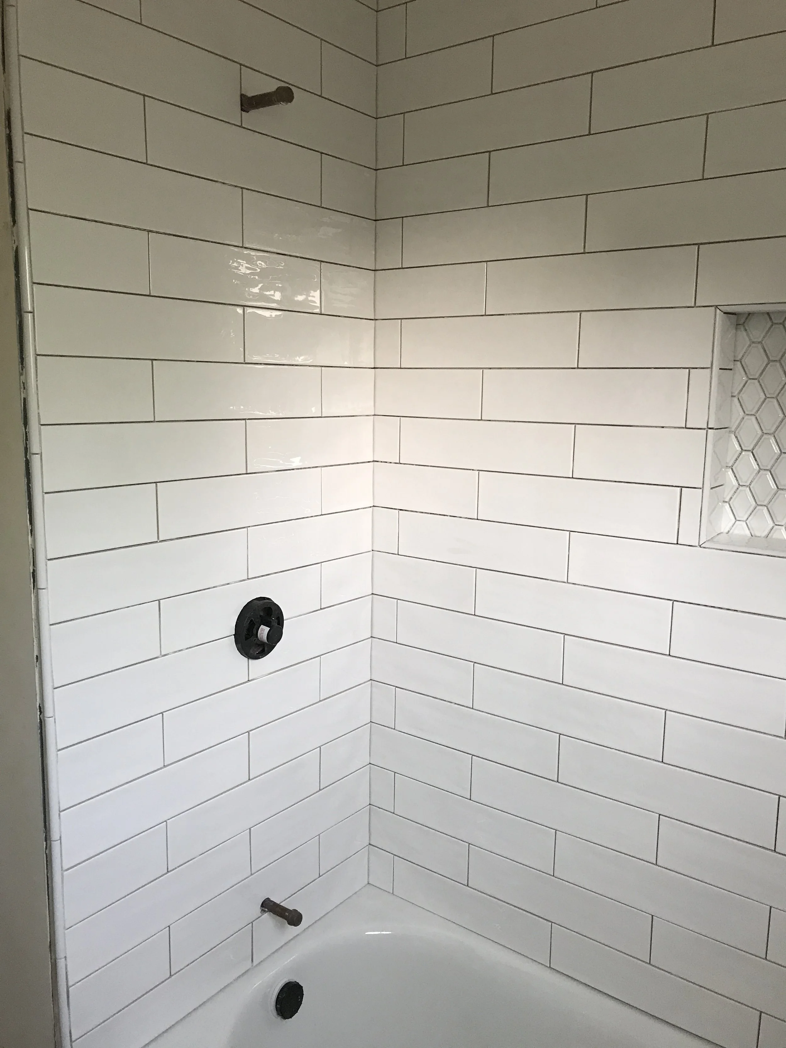 Inside a bathroom shower area with white subway tiles on the walls, a faucet hole and a drain cover visible, and a small built-in niche with hexagonal tiles.