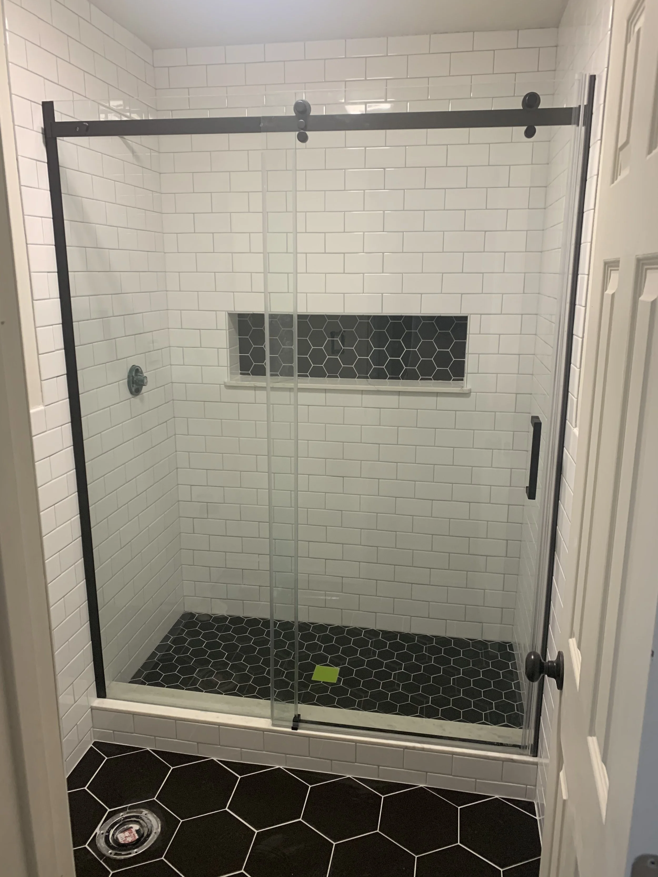 A modern shower with white subway tile walls, black hexagon floor tiles, a black-and-clear glass door, and a small built-in black tile niche.