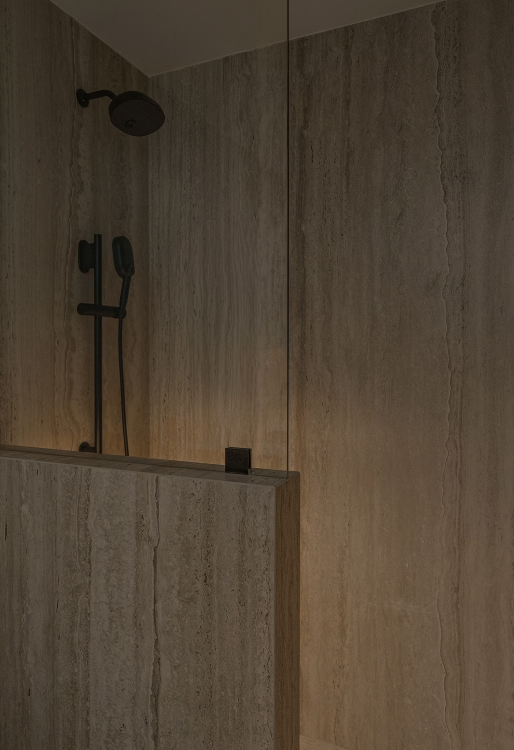 A shower area with a black showerhead, handheld shower, and wall-mounted control, set against textured beige stone tiles with a partially mirrored wall.