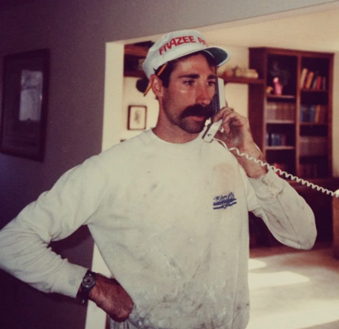 A man with a mustache wearing a white cap with red text, a white long-sleeve shirt, and a wristwatch, talking on a corded telephone inside a house.