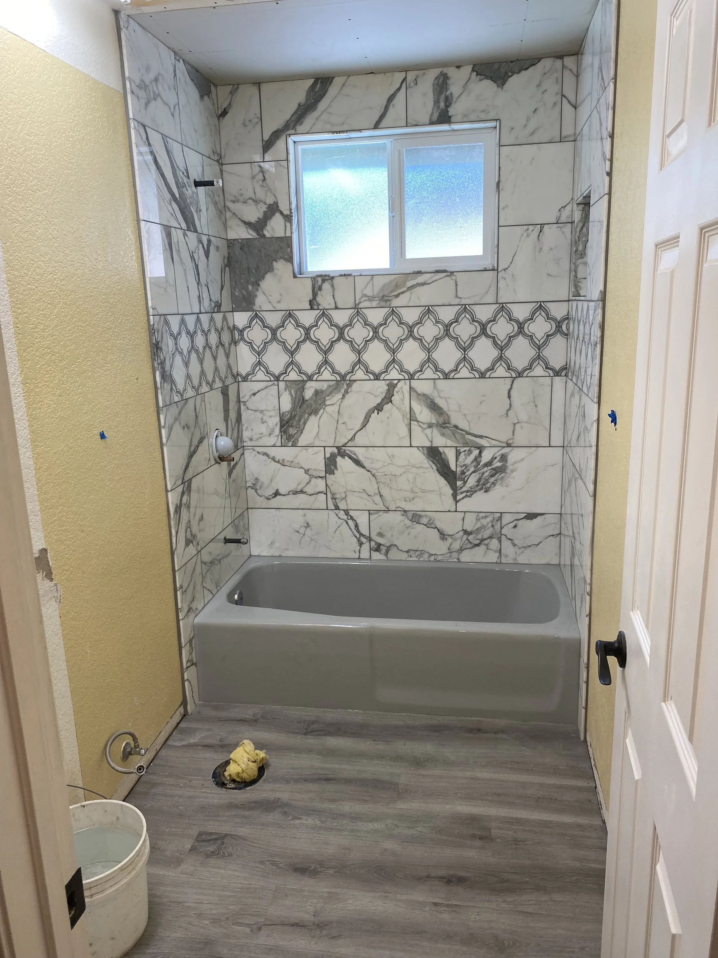 Bathroom with a bathtub, partially tiled walls with marble and patterned tiles, window, and flooring, during renovation or cleaning.