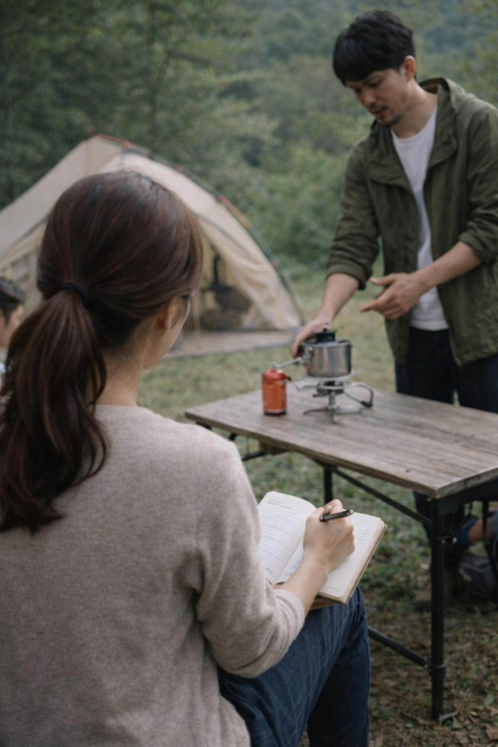 キャンプ場で女性がノートに書き込み、男性がガスコンロで調理をしている場面