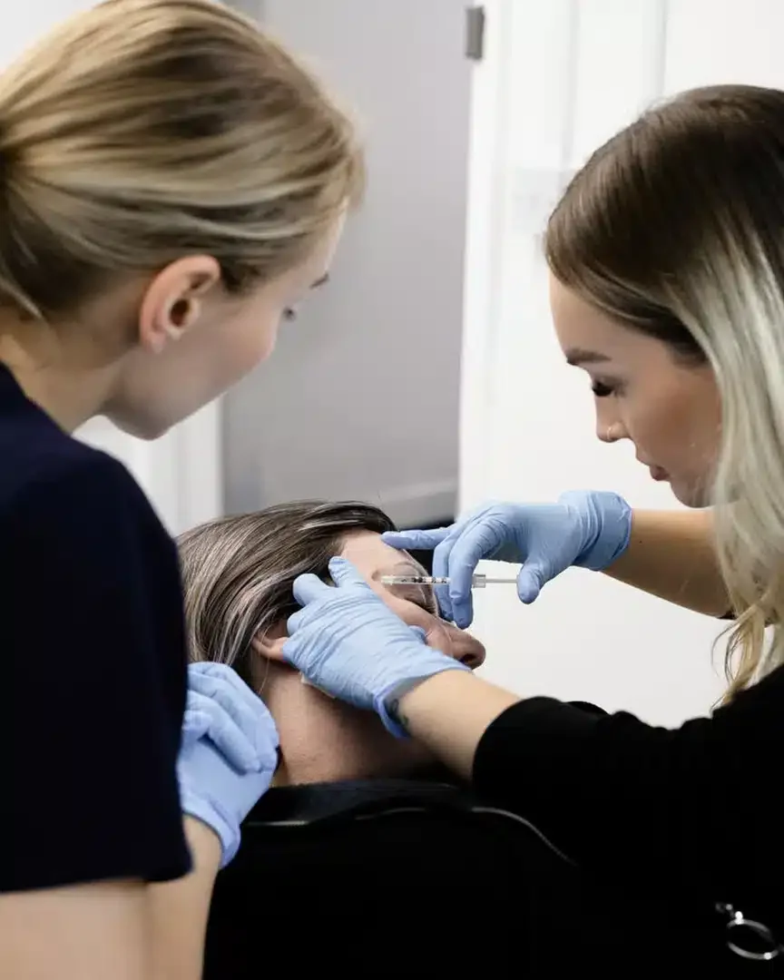 Two women wearing blue gloves administering a facial injection to a woman lying back with her eyes closed in a clinical setting.