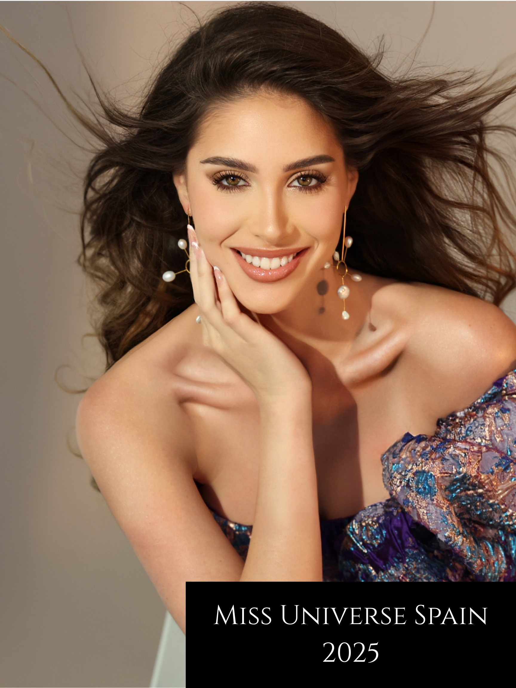 A smiling woman with long, flowing brown hair and light makeup, wearing elegant earrings with pearls and a colorful off-the-shoulder dress, promoting Miss Universe Spain 2025.