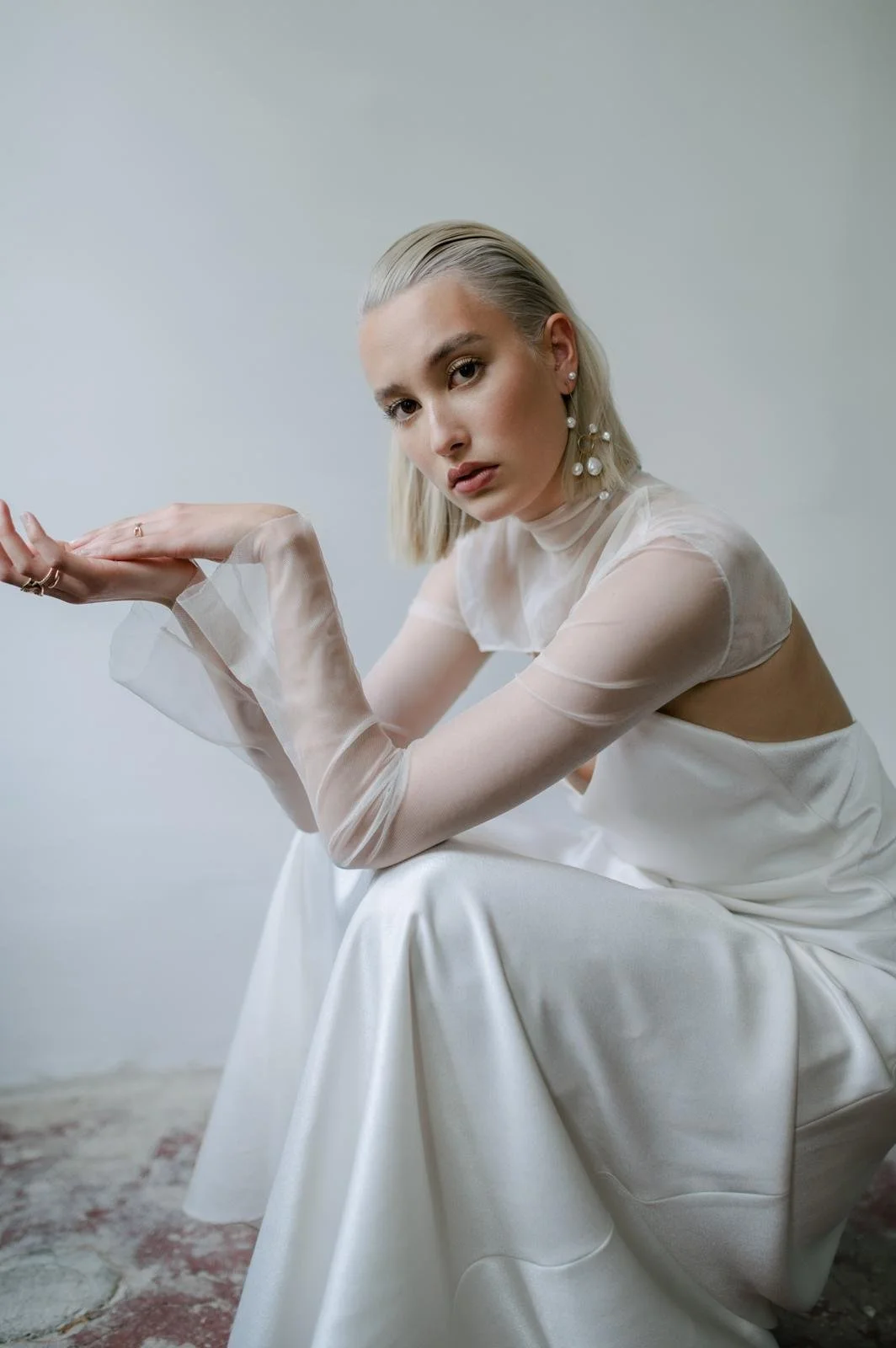 A young woman with blonde hair wearing a white satin dress with sheer, long sleeves and pearl earrings, posing against a plain gray background.