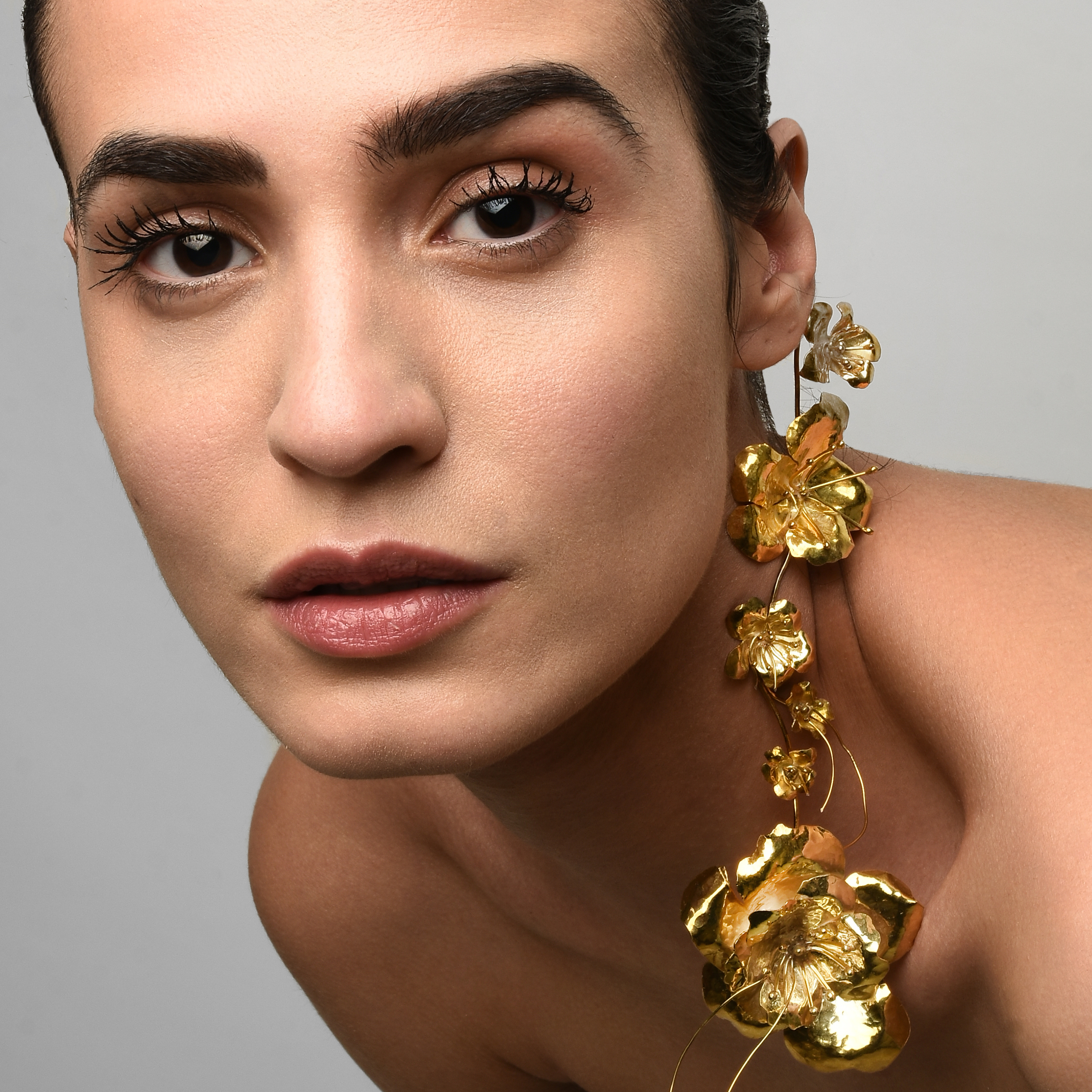 Close-up portrait of a woman with dark hair, wearing a large gold floral statement earring, and subtle makeup.