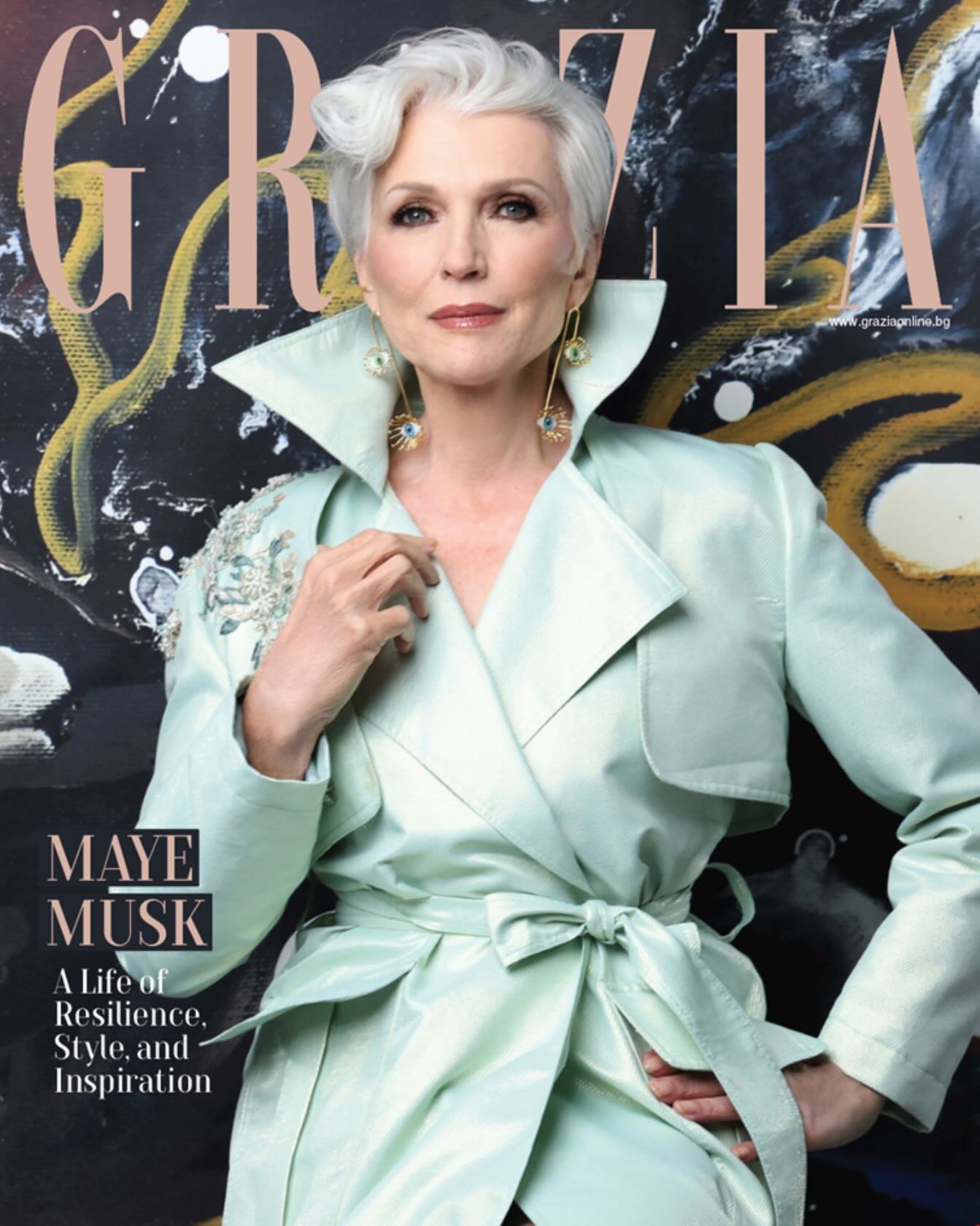 A woman with short, icy blonde hair wearing a light green trench coat with embroidered shoulders, large earrings, and a confident expression, standing in front of a black background with gold and white abstract designs, and the word 'GRAZIA' in large