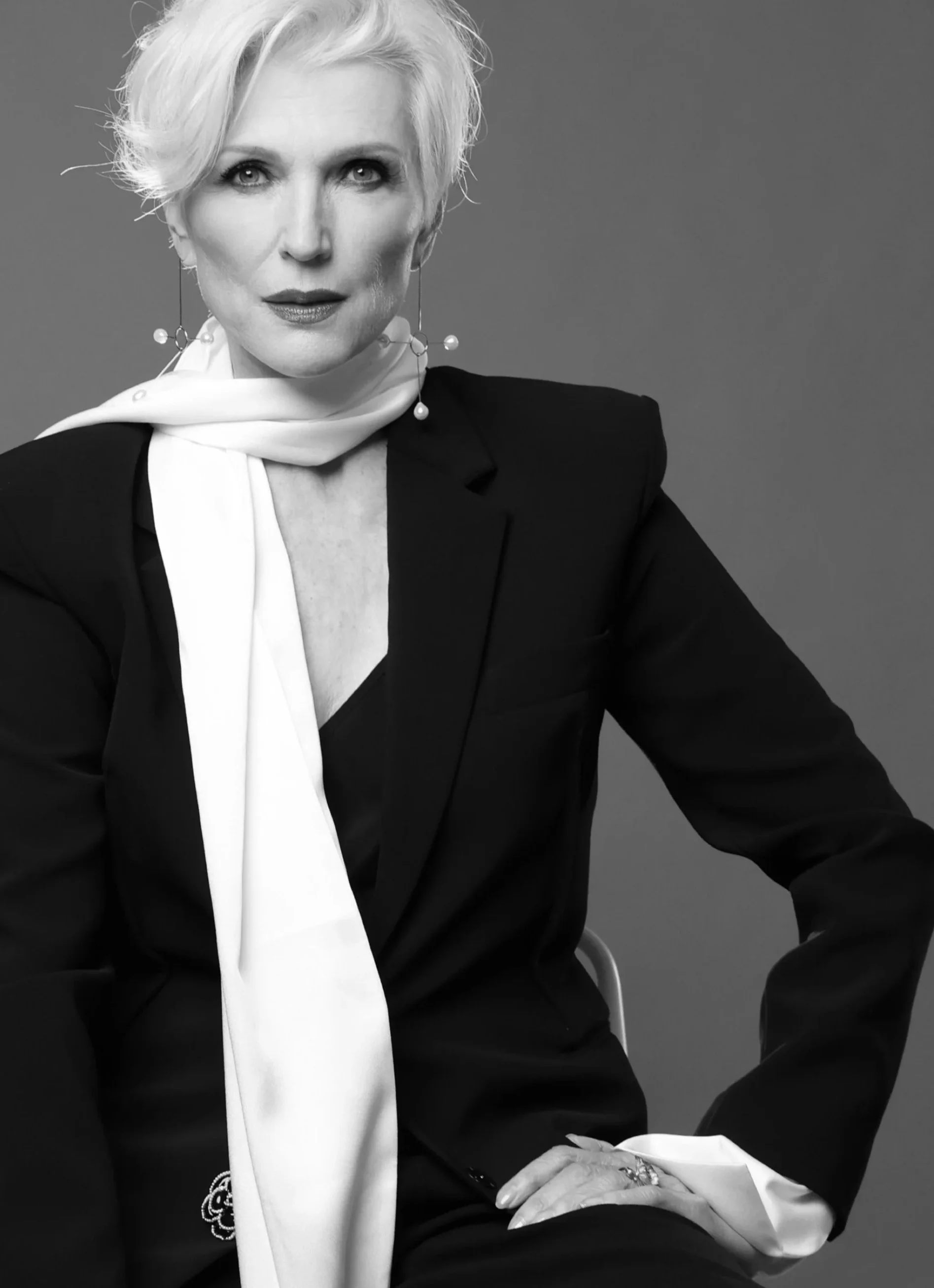 Black and white portrait of a woman with short, styled blonde hair, dressed in a black blazer, with a white scarf around her neck, gazing at the camera with a confident expression.