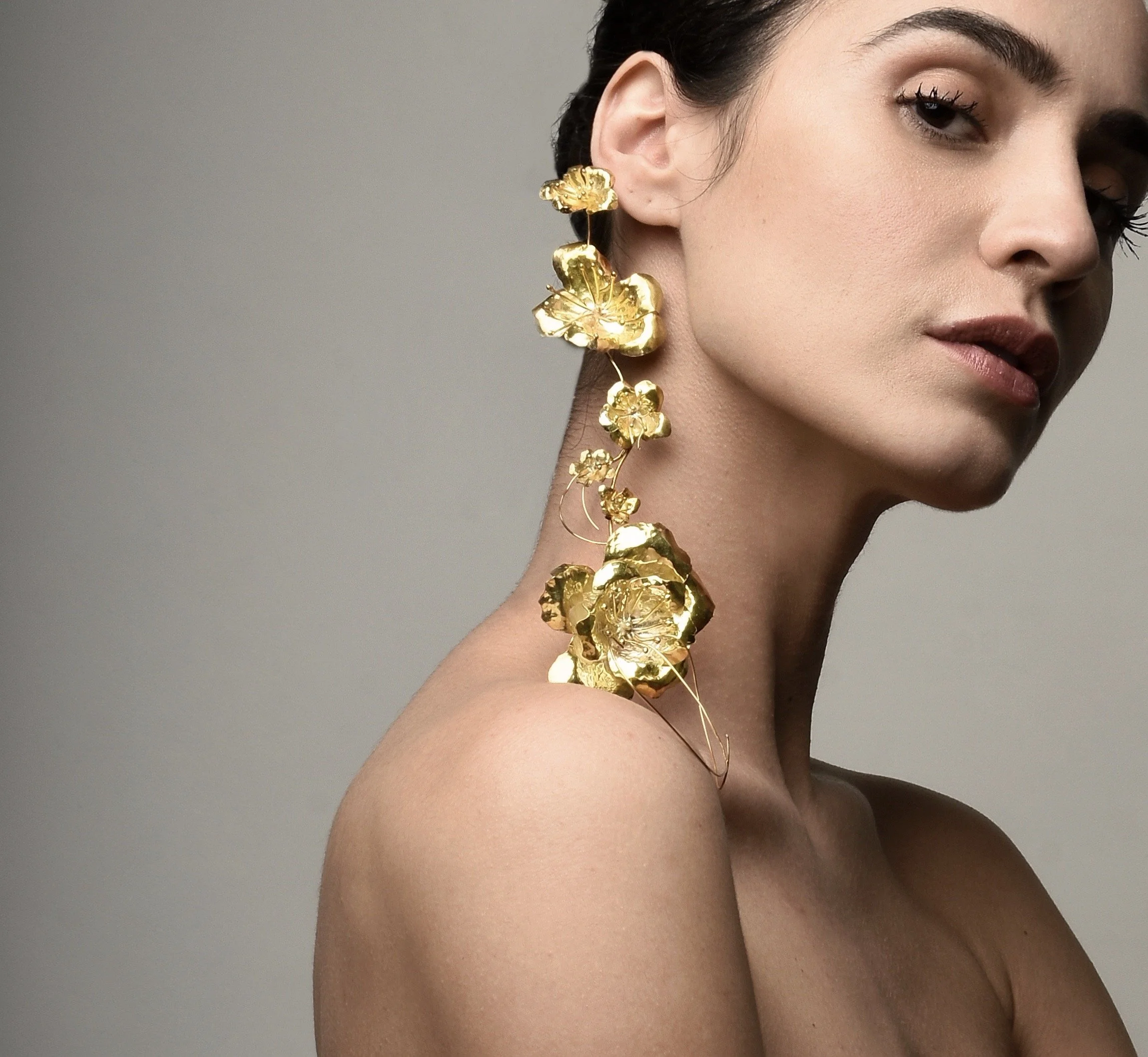 A woman with dark hair and light skin wearing large, intricate gold floral earrings.