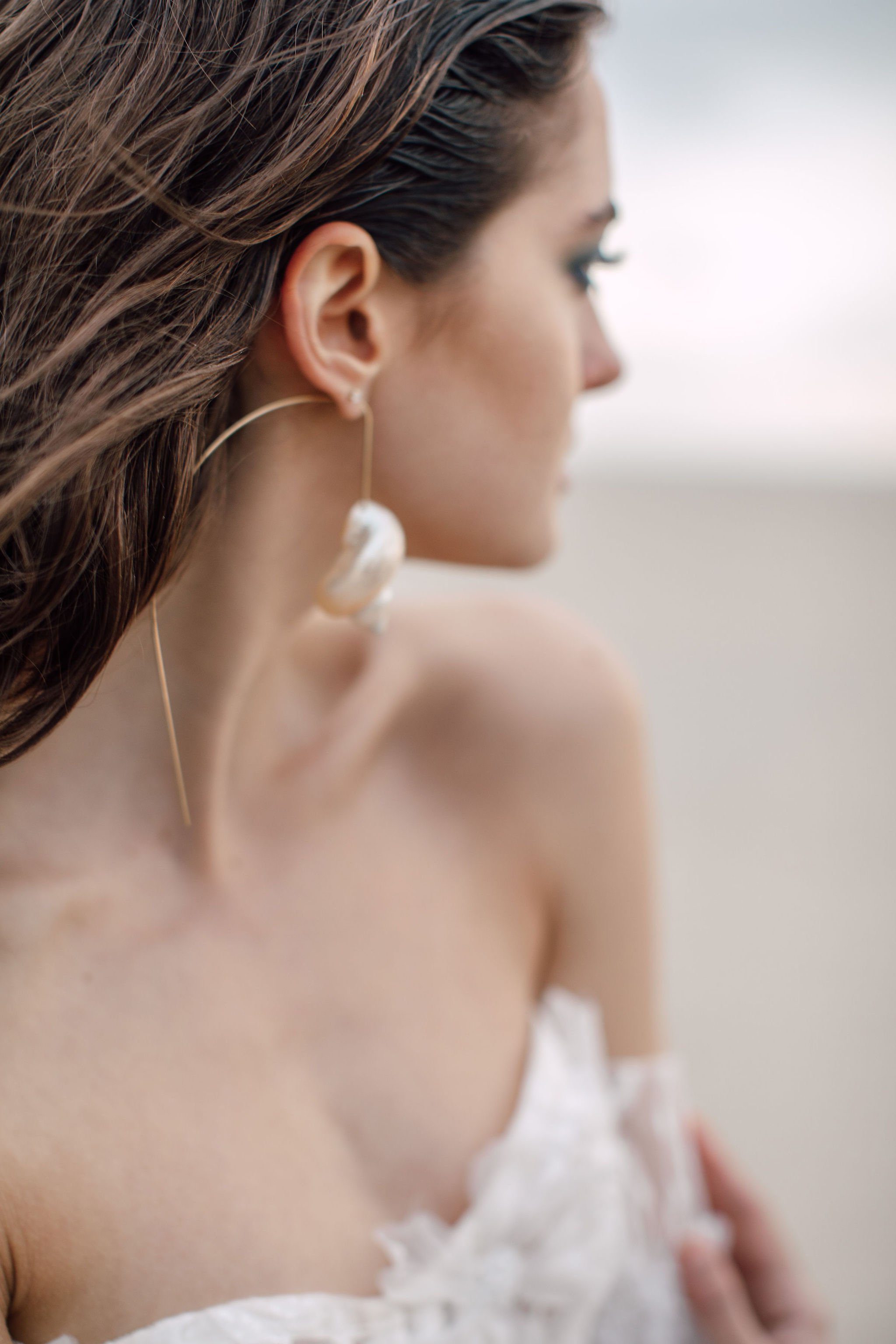 Close-up of a woman with long, brown hair wearing large, white shell earrings and a white off-the-shoulder top, looking to the side against a blurred background.