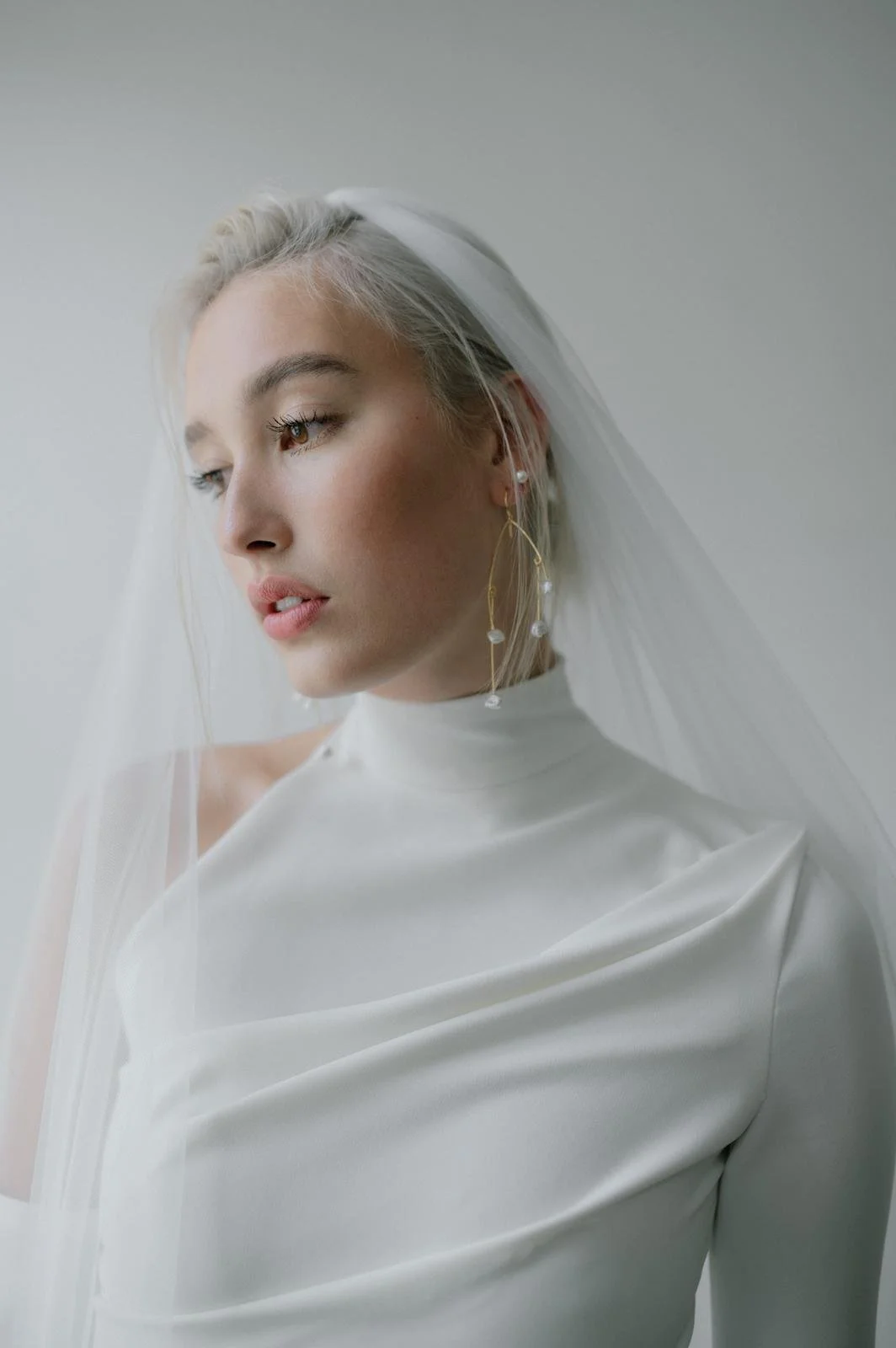 A woman with platinum blonde hair wearing a white dress and veil, accessorized with dangling earrings, looking down and to the side against a plain background.
