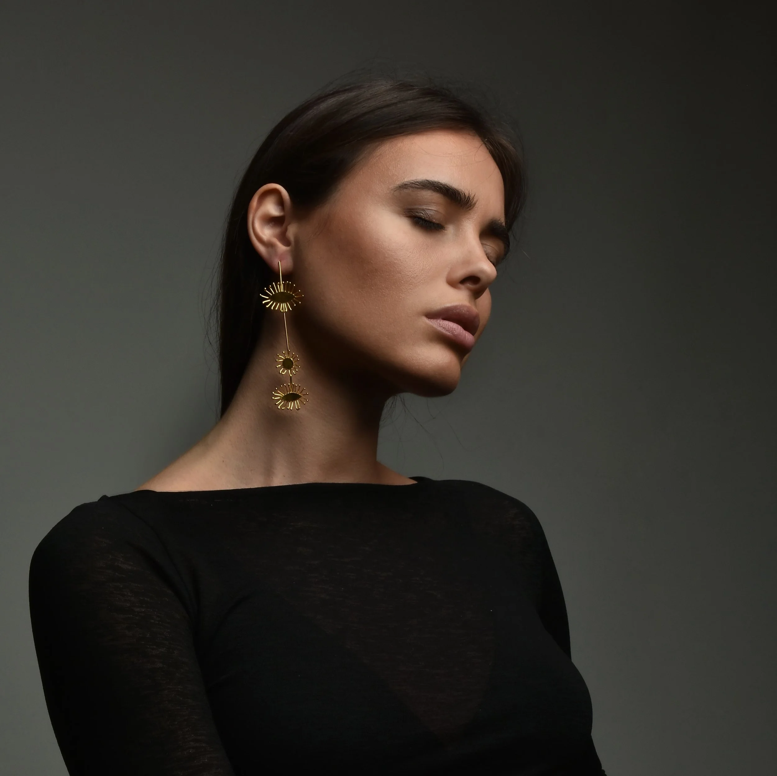 A woman with dark hair and earrings posing with her eyes closed against a dark background.
