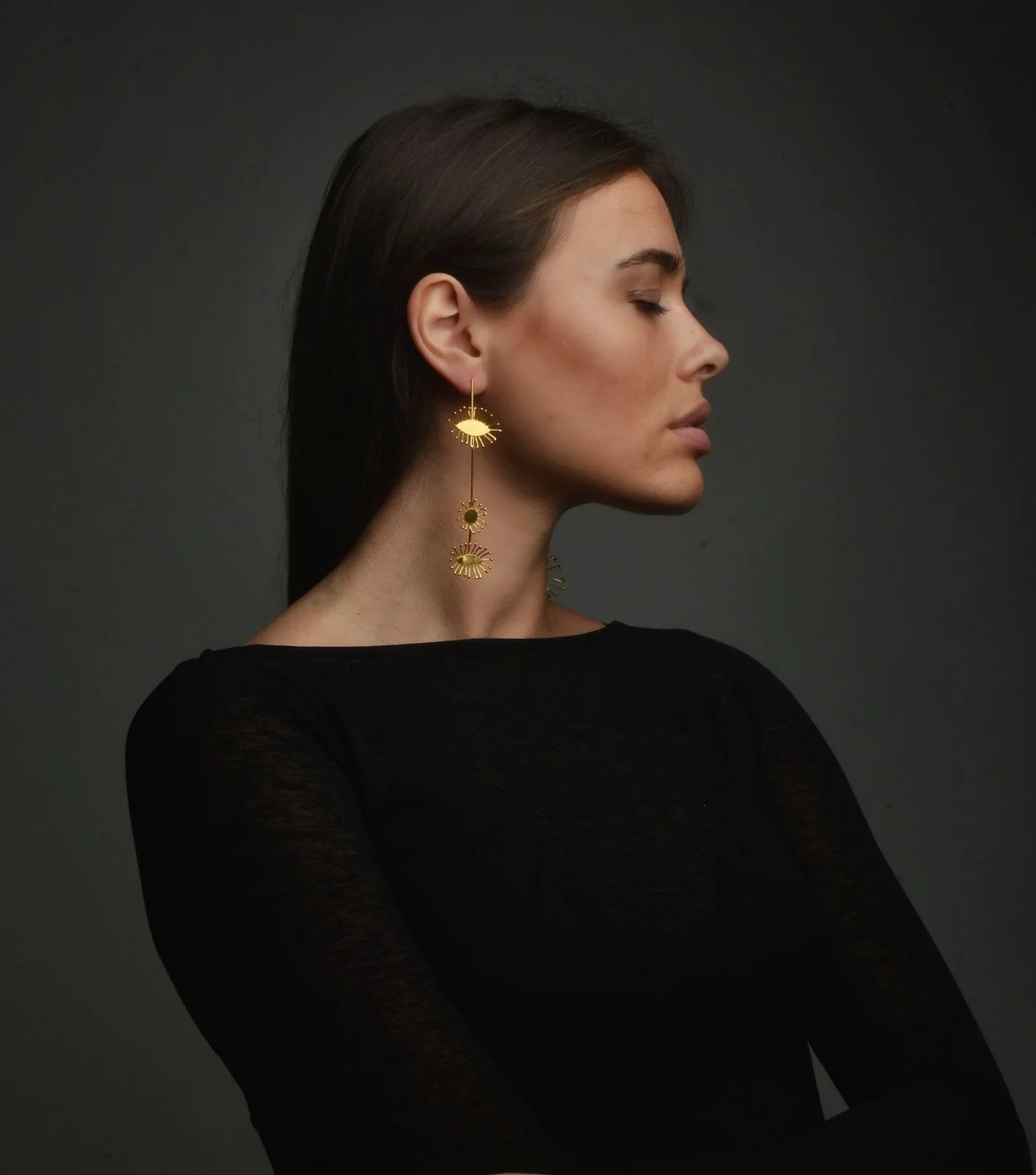 A woman with dark hair in profile wearing black top and gold eye-shaped earrings with sun motifs, against a dark background.