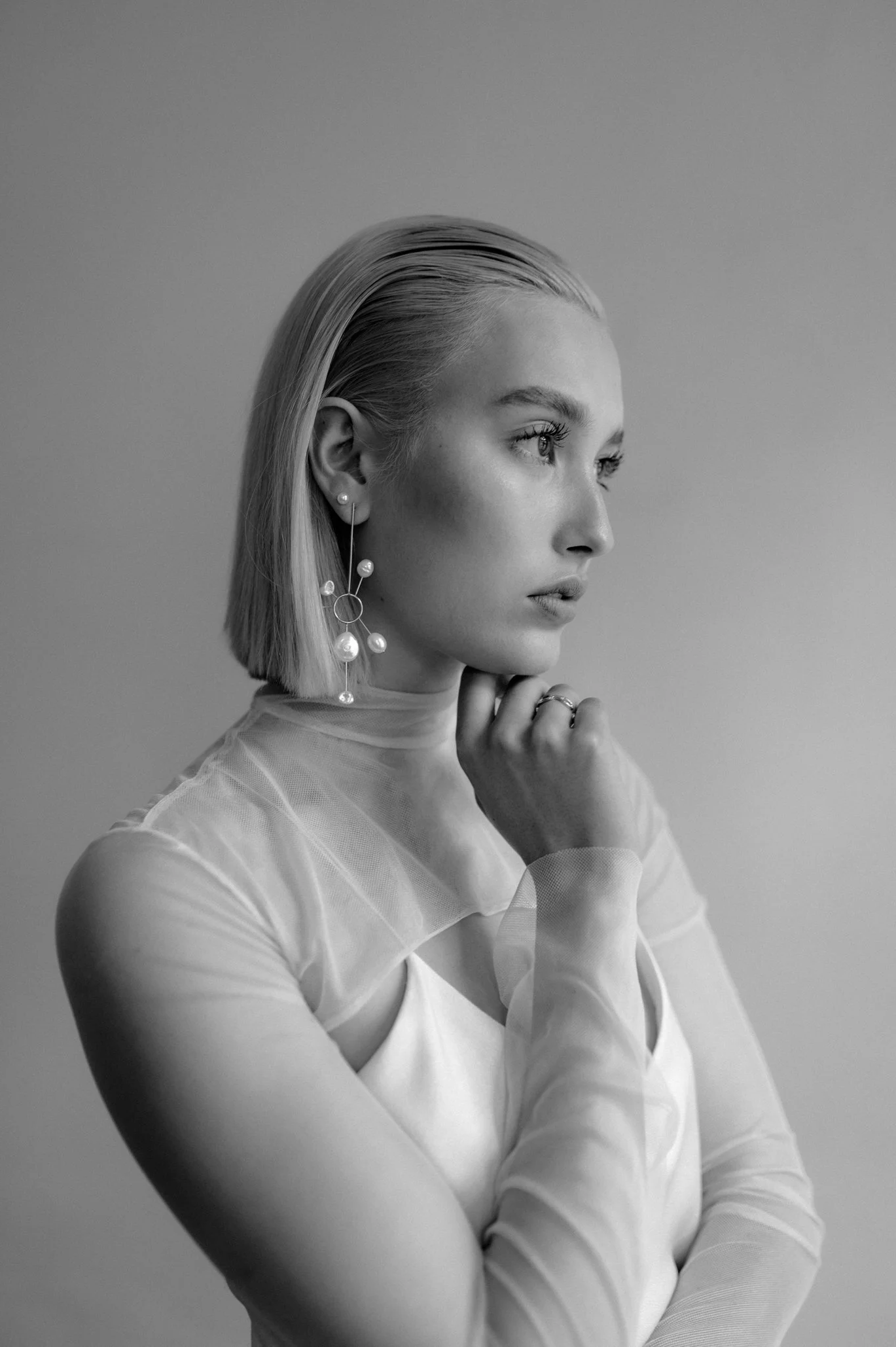 Black and white portrait of a woman with short, straight hair, wearing a sheer long-sleeve top and dangling pearl earrings, looking to her right with her hand resting near her chin.