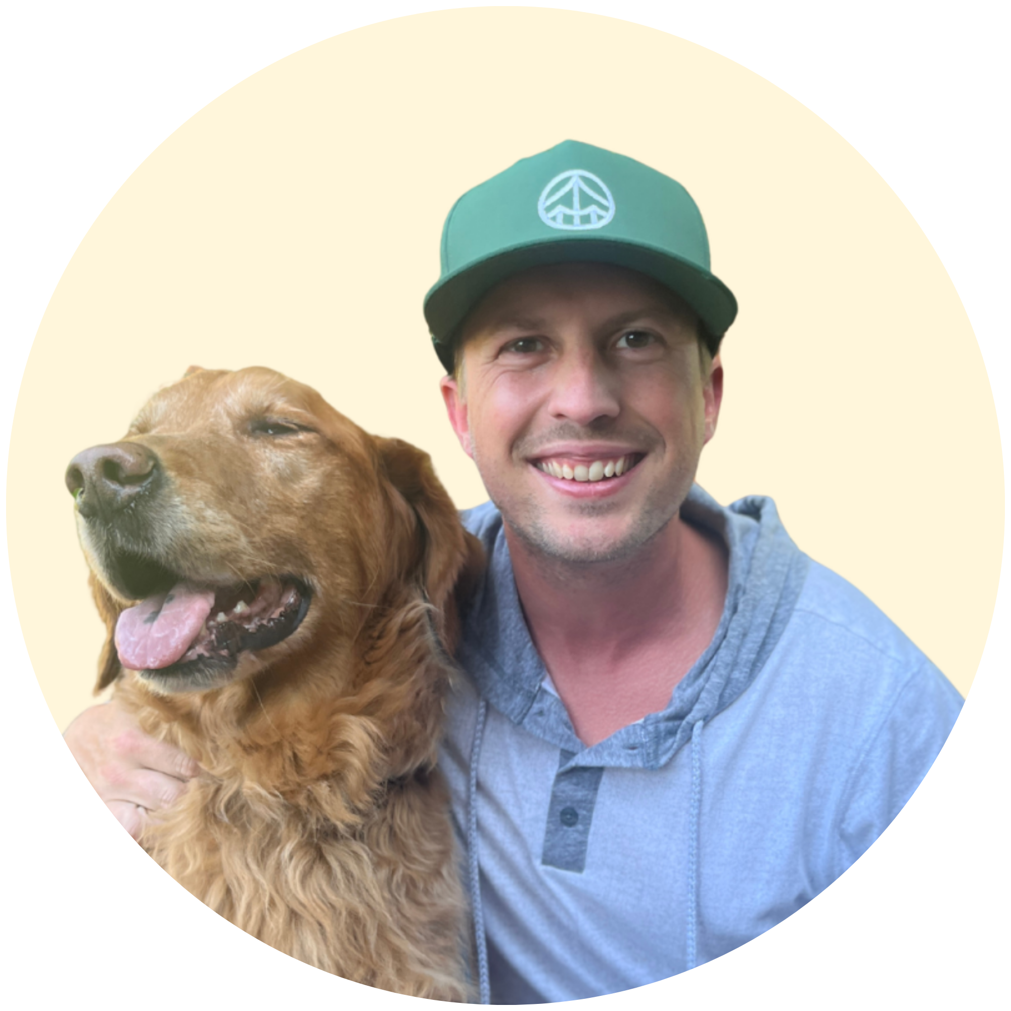 A smiling man in a blue denim shirt holding a golden retriever dog. The man is wearing a green baseball cap with a peace sign logo. The dog appears relaxed and happy.