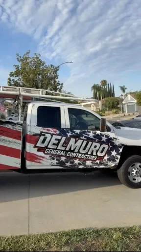 White company truck with Del Muro General Contractors logo, parked in a residential neighborhood with trees, a house, and a blue sky with clouds.