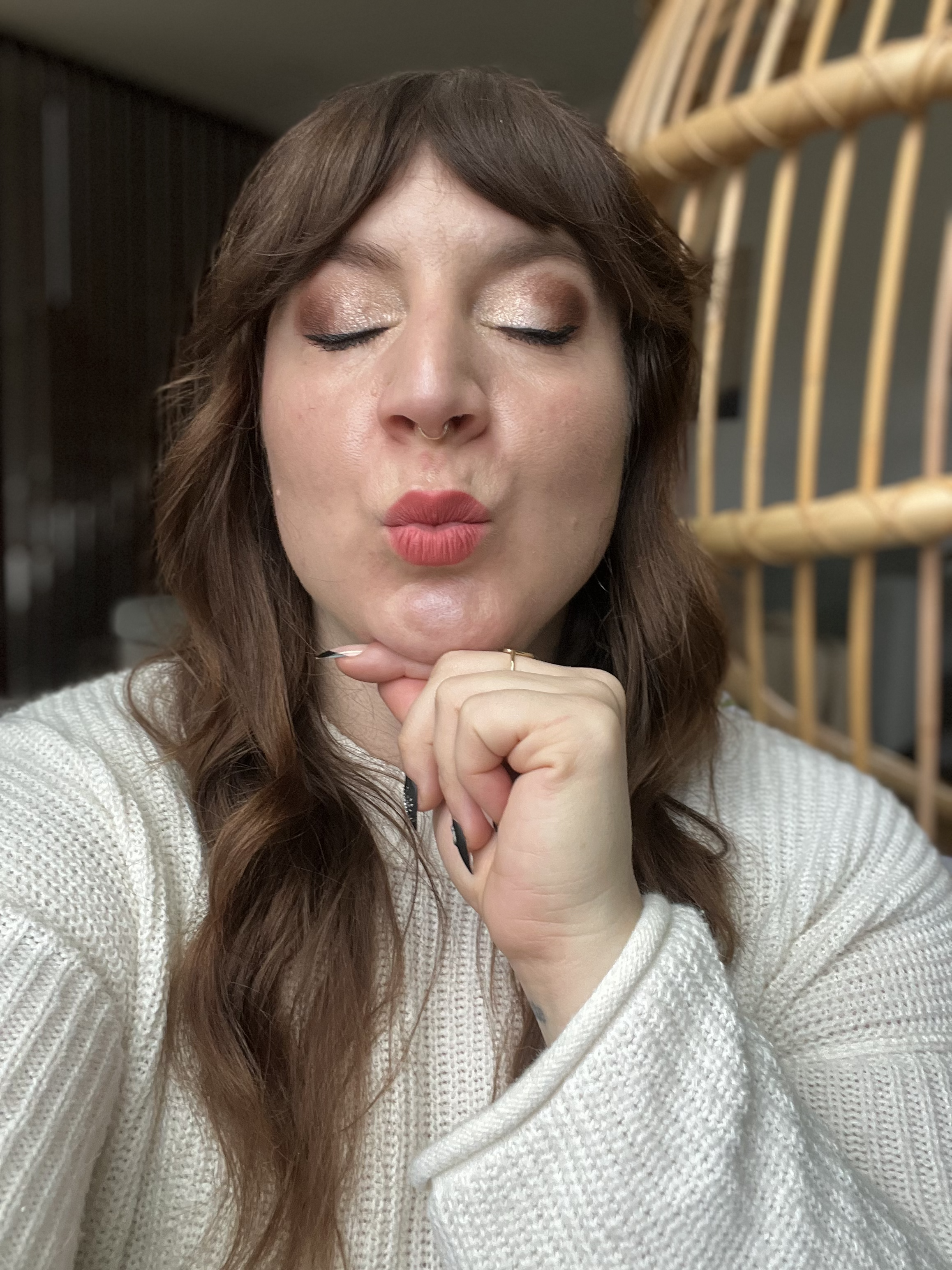 A woman with closed eyes and pursed lips, making a kiss face, rests her chin on her hand with a wicker headboard in the background.