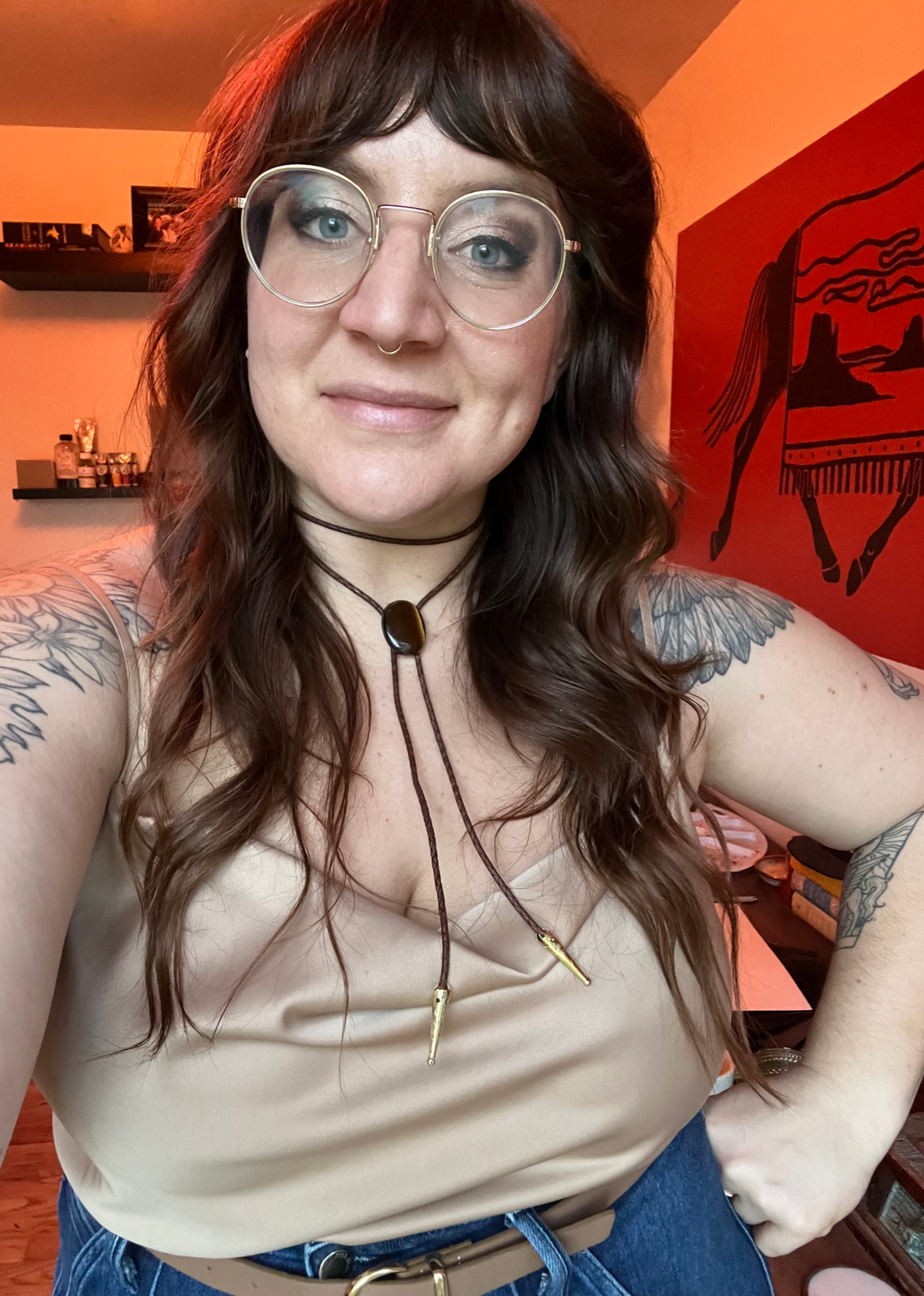 A woman with long wavy brown hair and glasses takes a selfie indoors. She has tattoos on her arms, a septum piercing, and is wearing a beige sleeveless top with necklaces. There is a red wall art of a jaguar behind her.