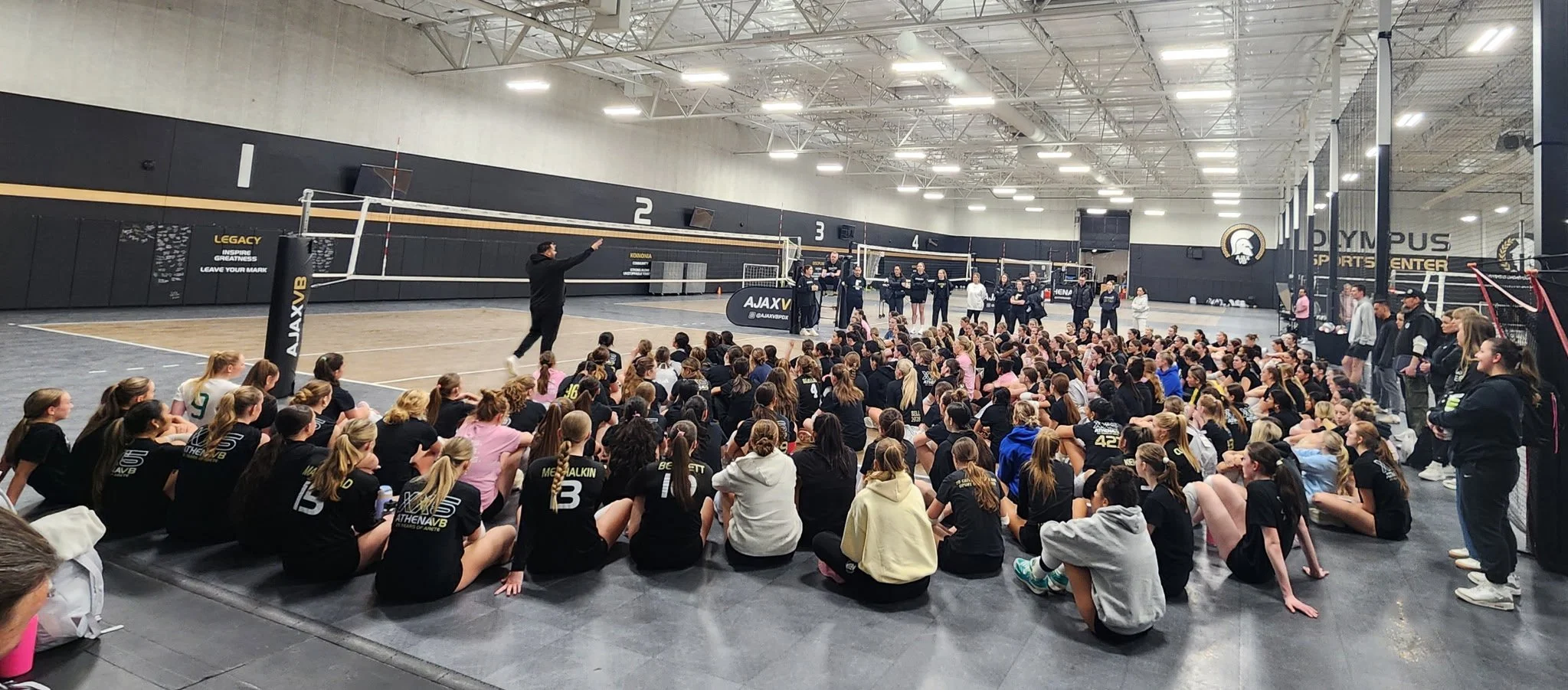 Group of youth and high school female and male volleyball players seated on the gym floor, listening to a coach, in a large indoor sports complex with volleyball courts and spectators watching. The facility is called Olympus sport center.