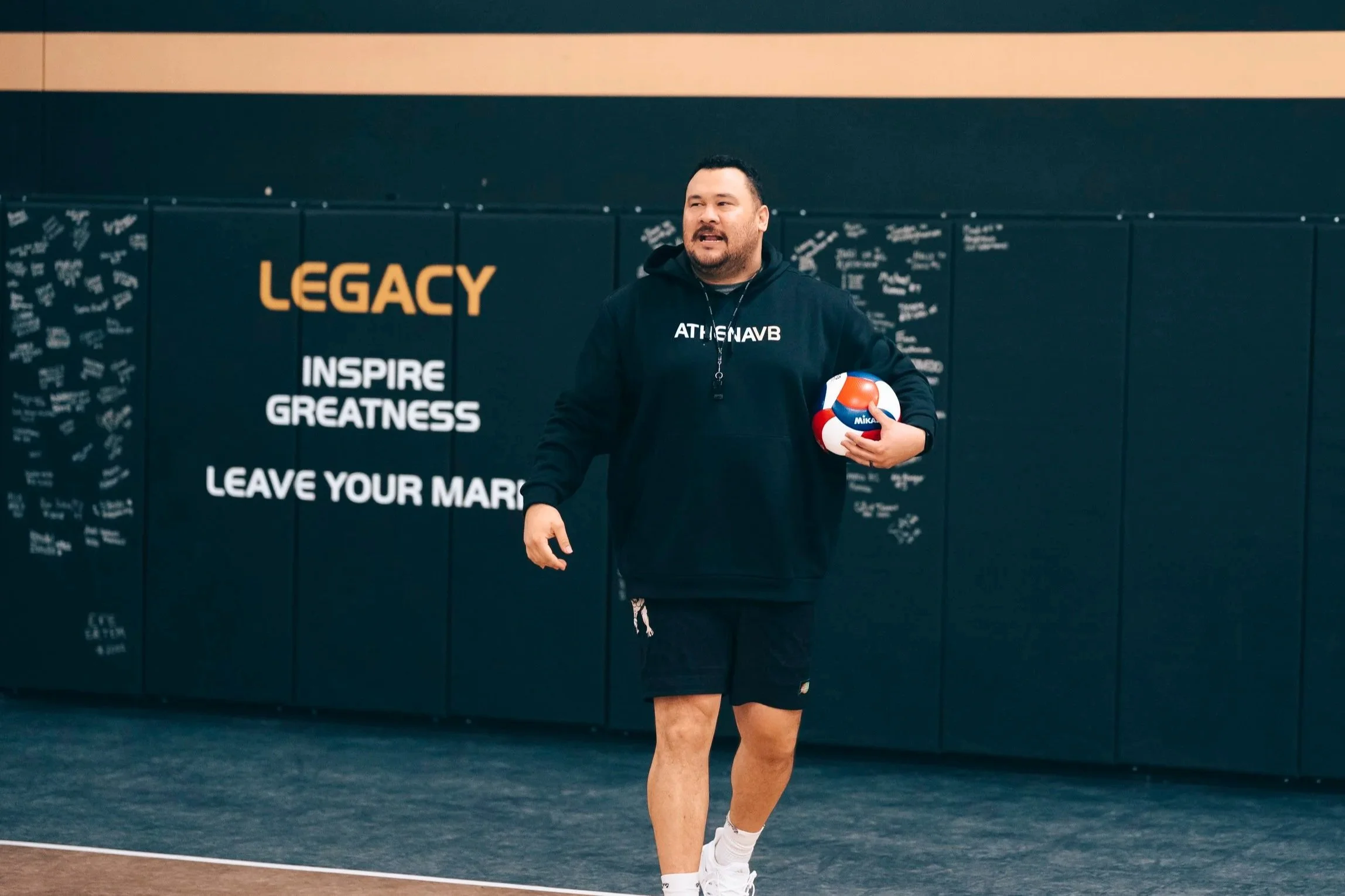 Coachk808, A man in black adidas athletic clothing holding a red, white, and blue LOVB volleyball in a gym with the words 'LEGACY, INSPIRE GREATNESS, LEAVE YOUR MARK' on the wall behind him.