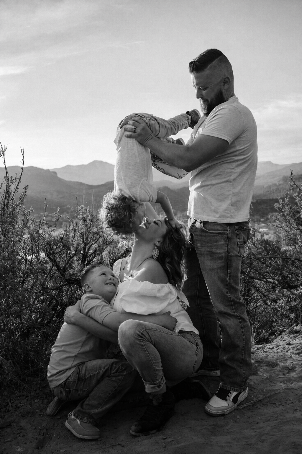 A family of four outdoors, with mountains in the background. The father is lifting a young girl upside down, and the mother is kneeling, holding a young boy. Everyone is smiling and enjoying the moment.