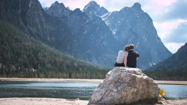 Two people sitting side by side on a rock near a still lake, framed by snow‑topped mountains and trees, creating a peaceful, supportive scene.