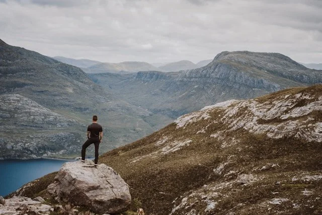 A solitary figure faces an expansive mountain range, standing steady on a rocky ledge as soft light moves across the terrain.
