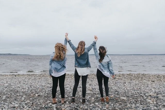 Three people standing on a rocky beach facing the ocean, with two raising their arms under an overcast sky.